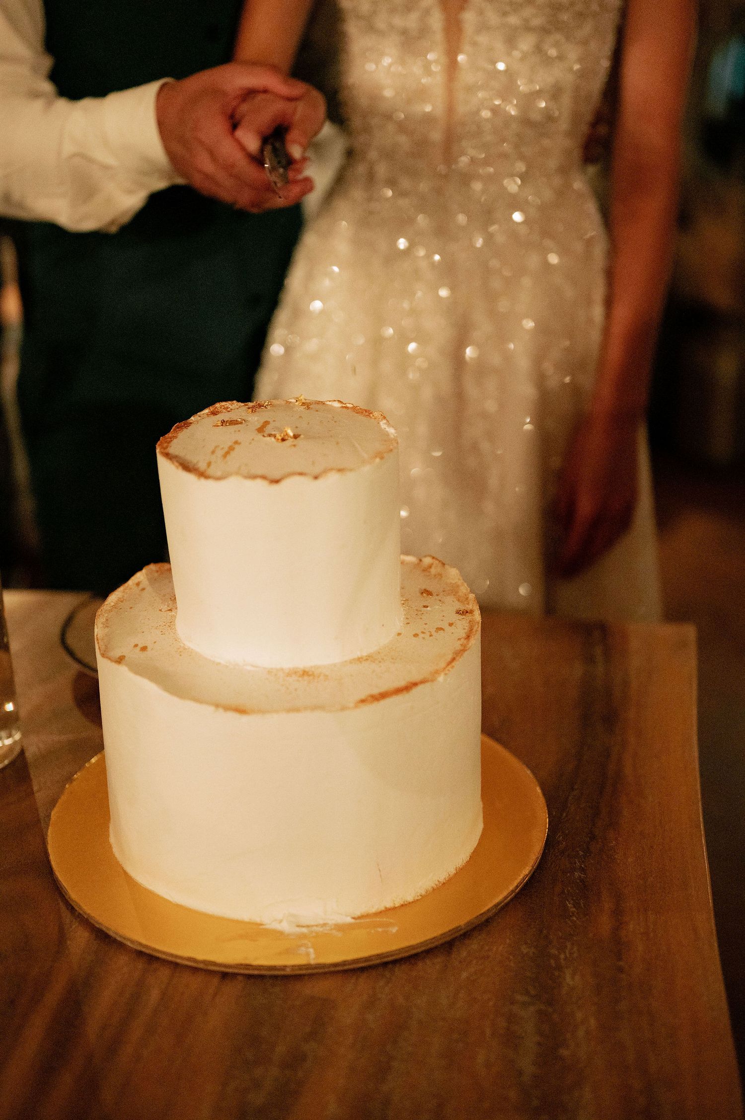 Simple white wedding cake displayed on wooden table with sparkly dress in background.