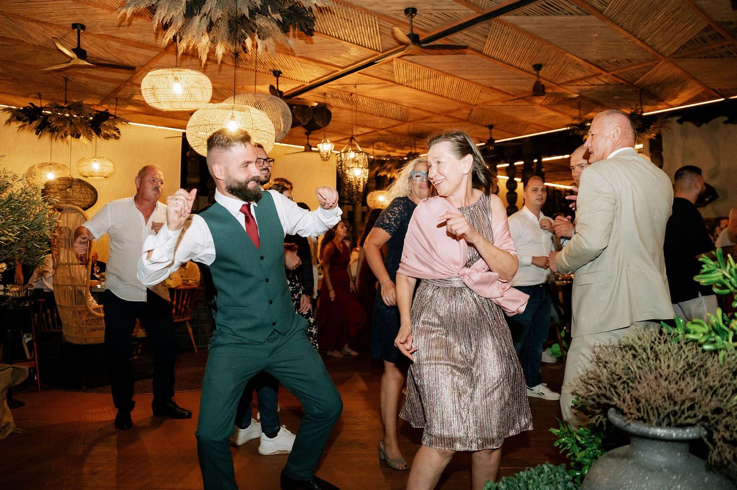 Wedding guests dancing enthusiastically at reception with wooden ceiling and ambient lighting.