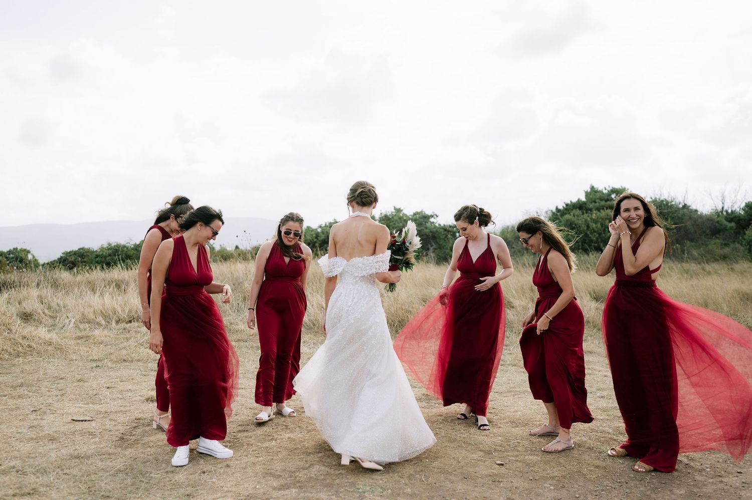 Bridesmaids in deep red dresses gather around bride in white on outdoor hillside.