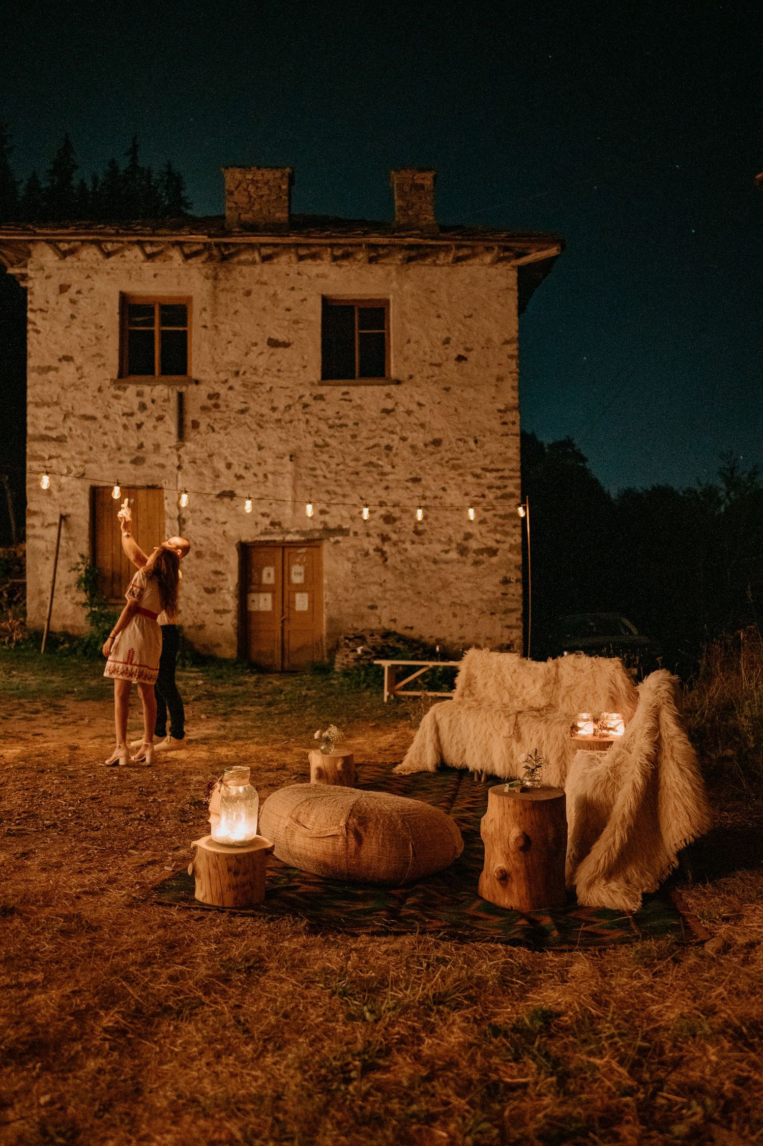Cozy outdoor seating area with string lights and fur throws outside a rustic stone house at night.