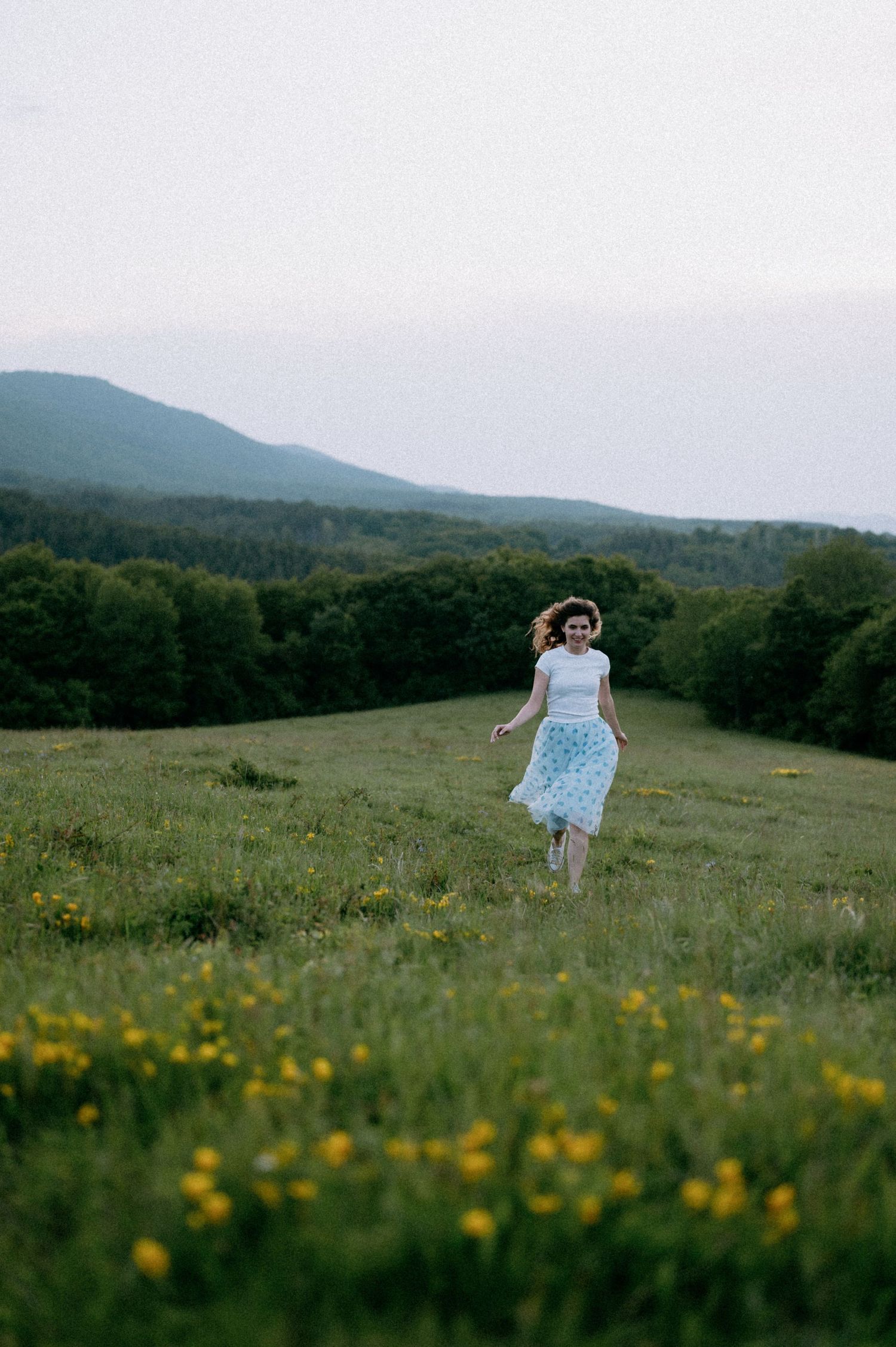 Person in flowing dress running through wildflower field with mountains in background.