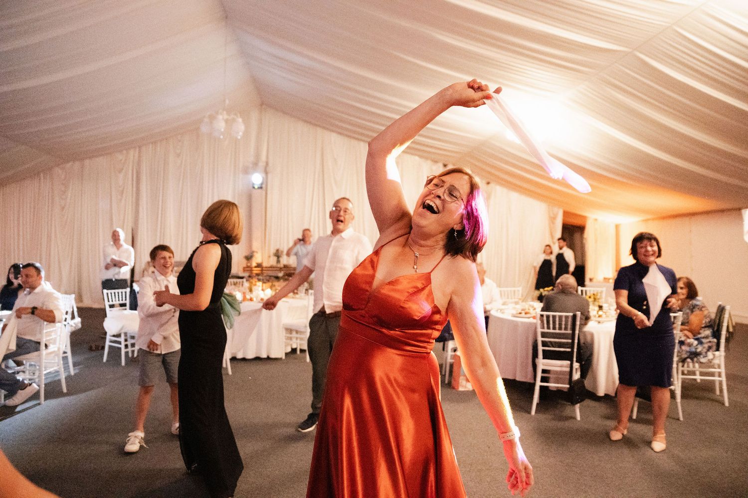 Wedding guests dance and celebrate on a dance floor under a white tent during a reception celebration.