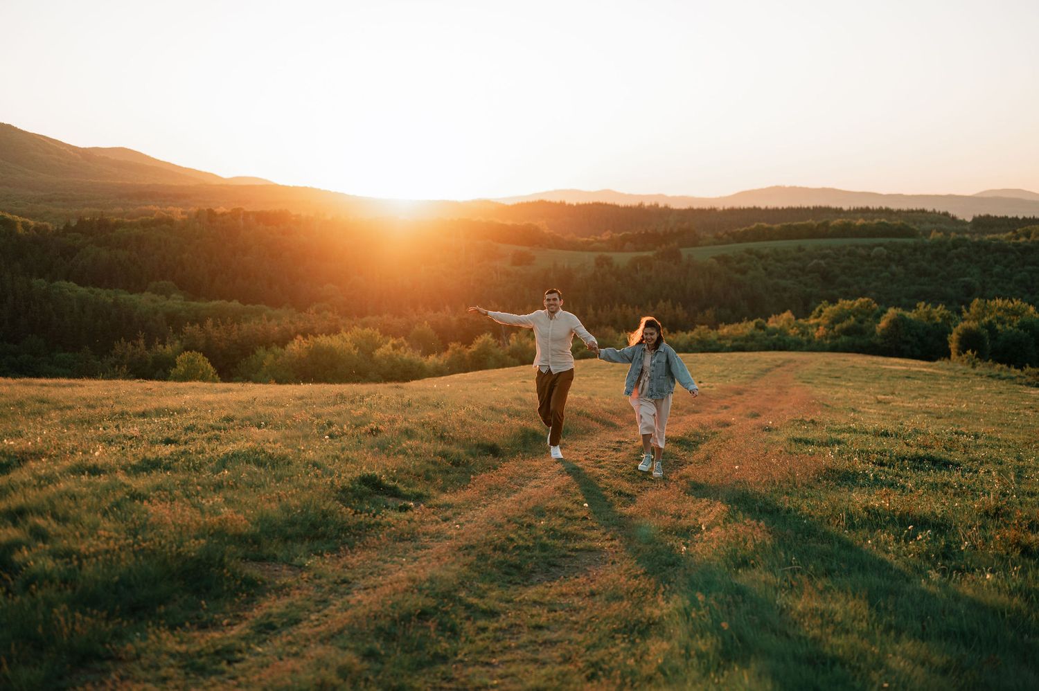 Silhouetted figures walk along a hilltop path at golden hour as the sun sets over rolling countryside meadows.