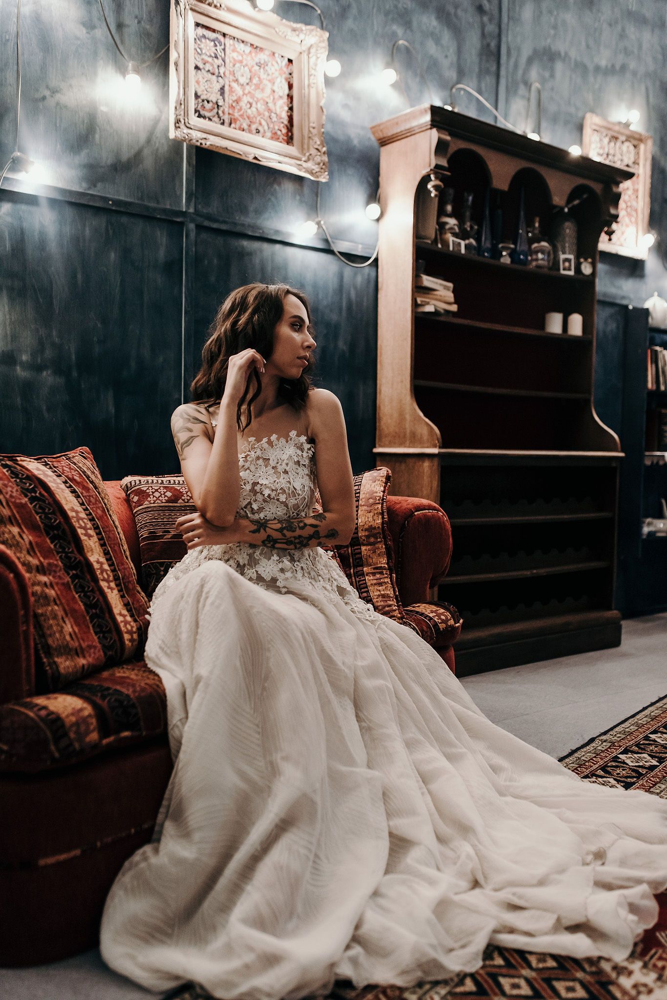 A bride in a flowing white wedding dress sits elegantly on a vintage couch against a dark wall.