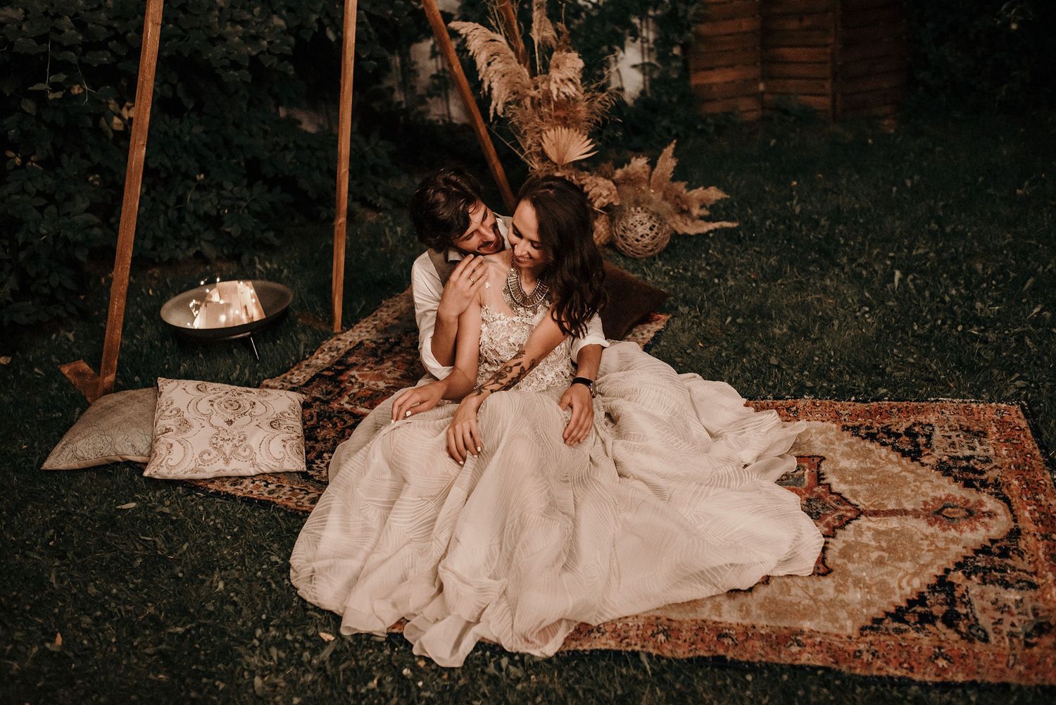 A vintage rug lies on dark grass with the flowing fabric of a white dress cascading across it in an outdoor setting.
