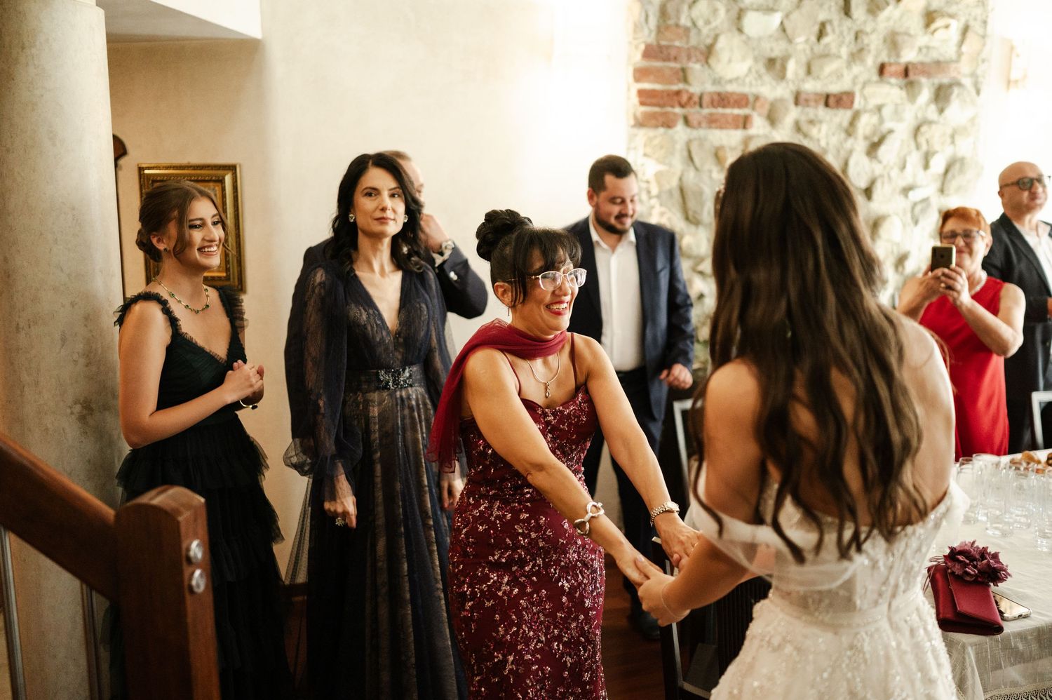 A group of wedding guests gather and socialize in a bright indoor space with stone walls.