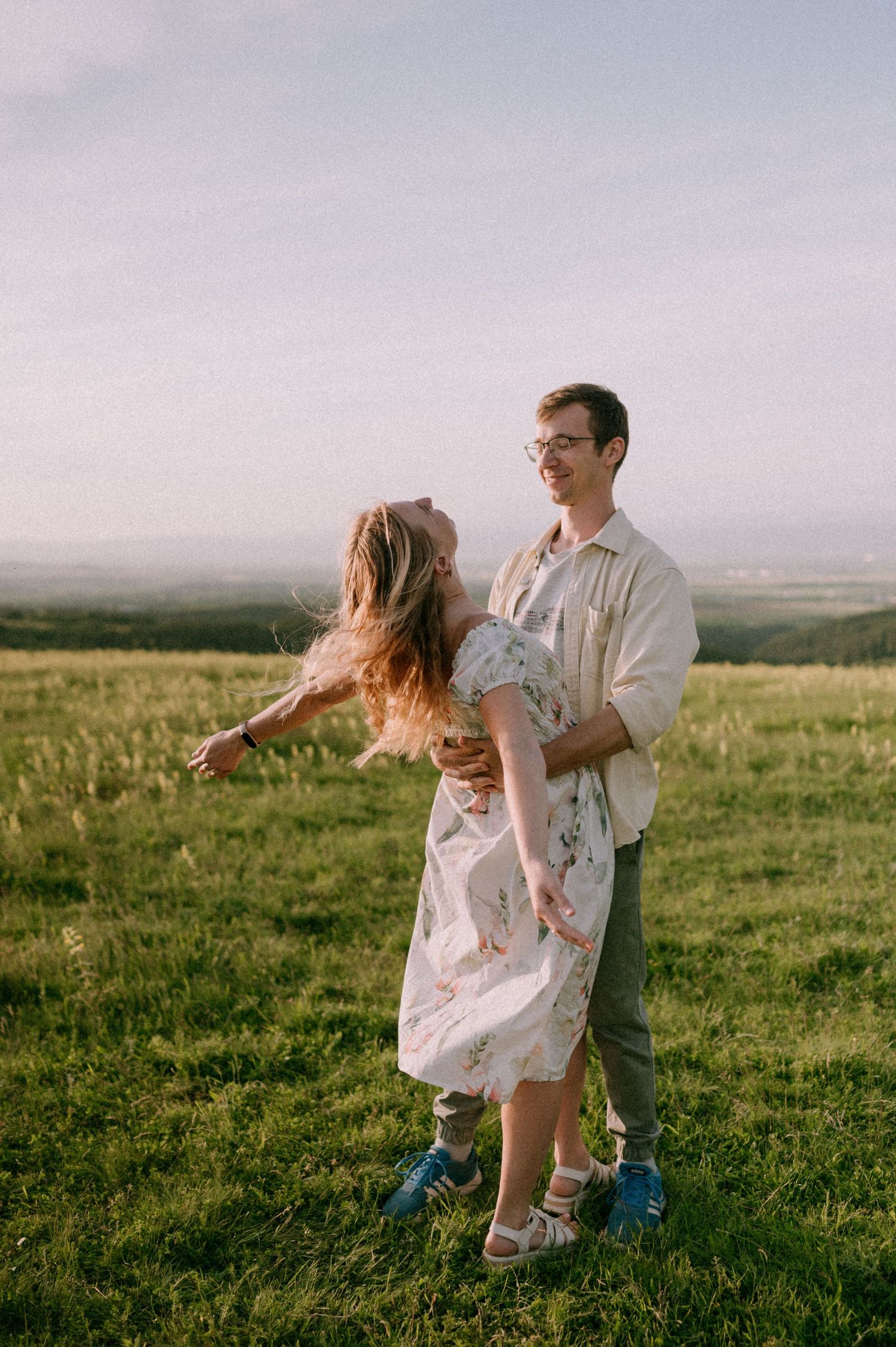 Playful couple dancing and spinning in a sunlit meadow with mountains in the background.