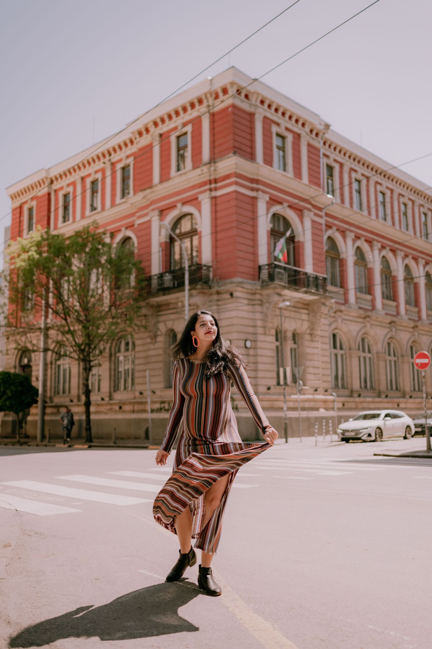 Woman in striped maxi dress posing on sunny street corner near ornate red historic building.