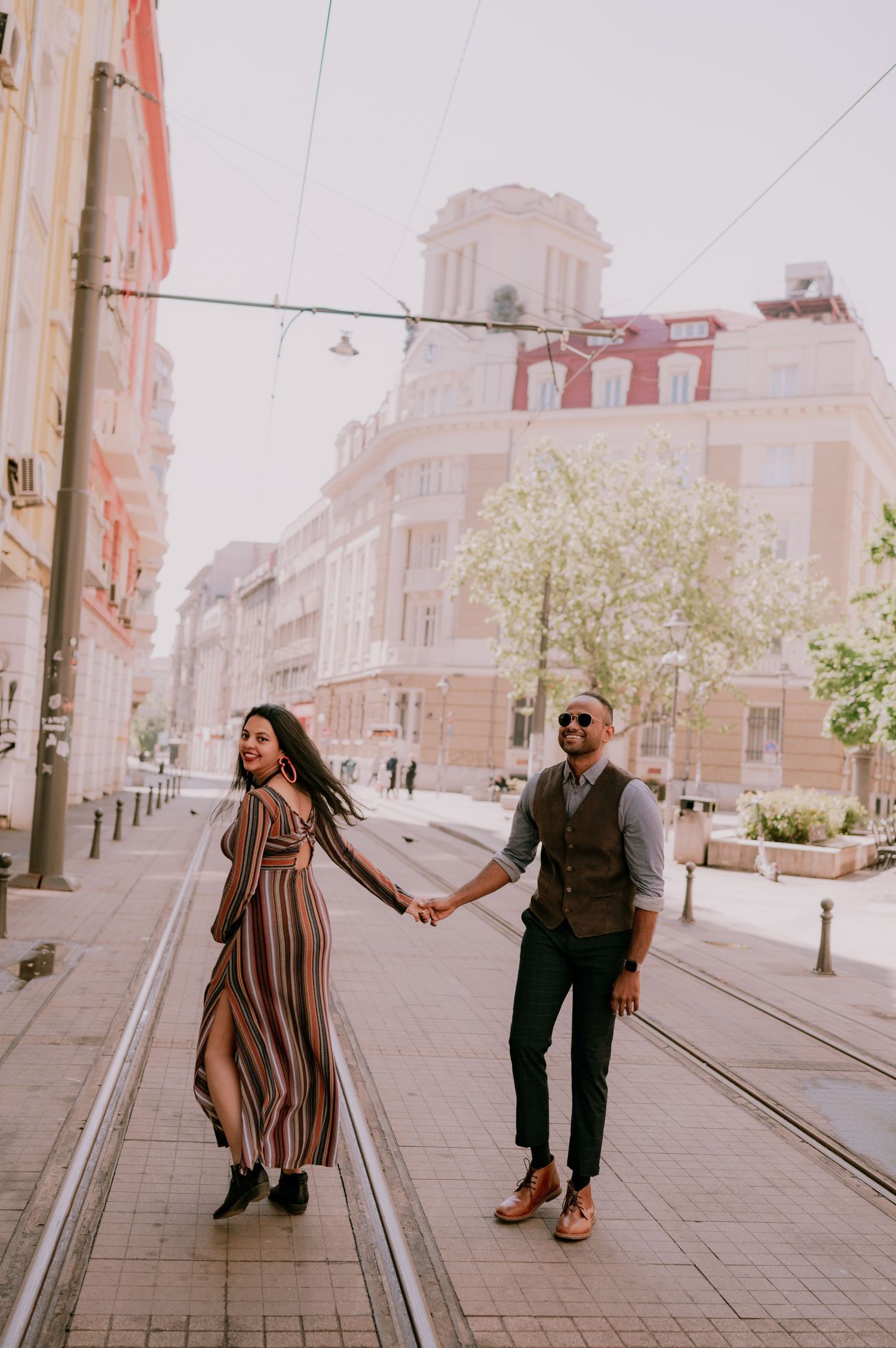 Couple holding hands walking on tram tracks in a vibrant city street, woman in striped dress, man in vest and sunglasses.