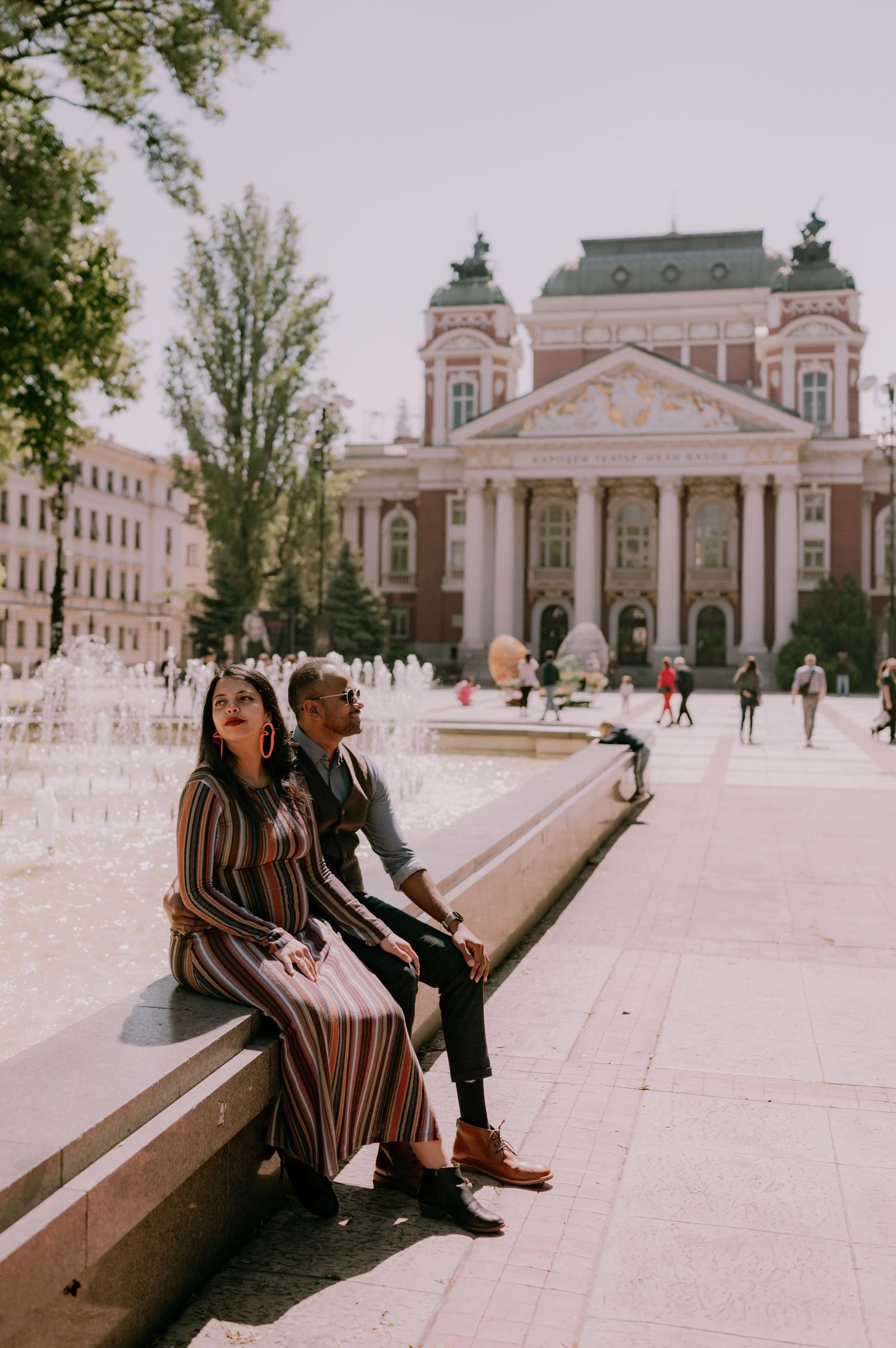 Couple sitting by a fountain in front of a grand neoclassical building in a sunny European city square.