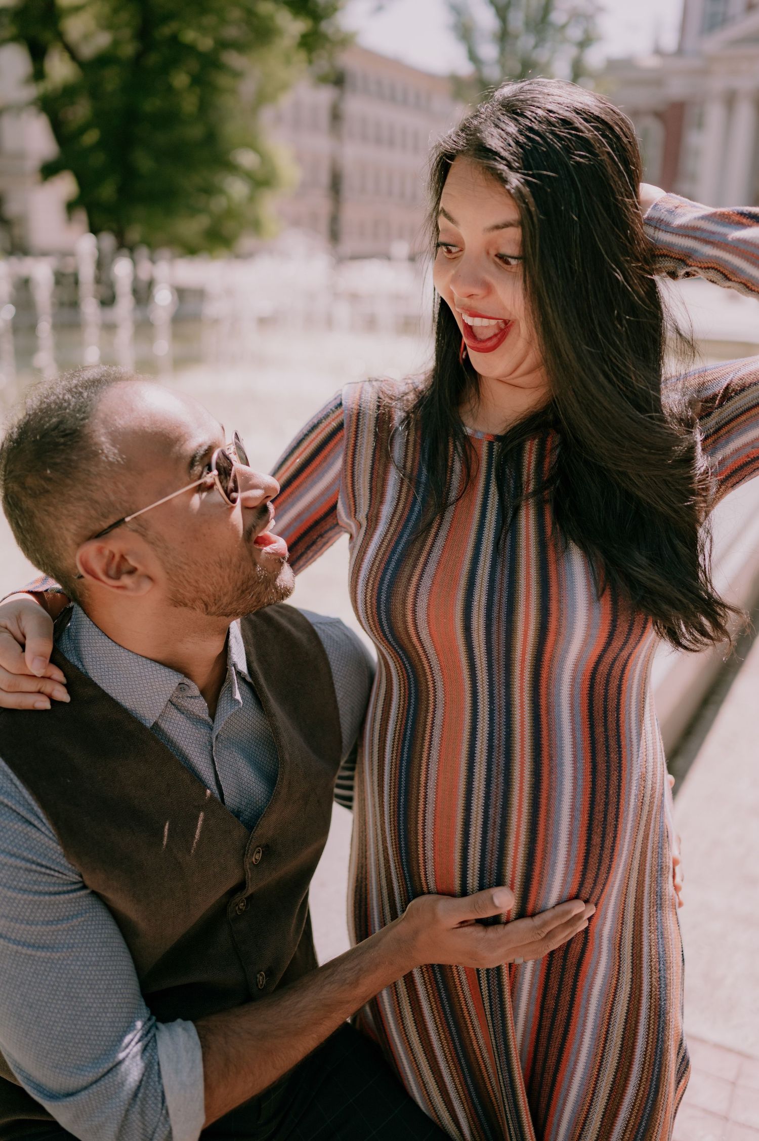 A couple shares a joyful moment outdoors, with the man tenderly touching the pregnant woman's belly.
