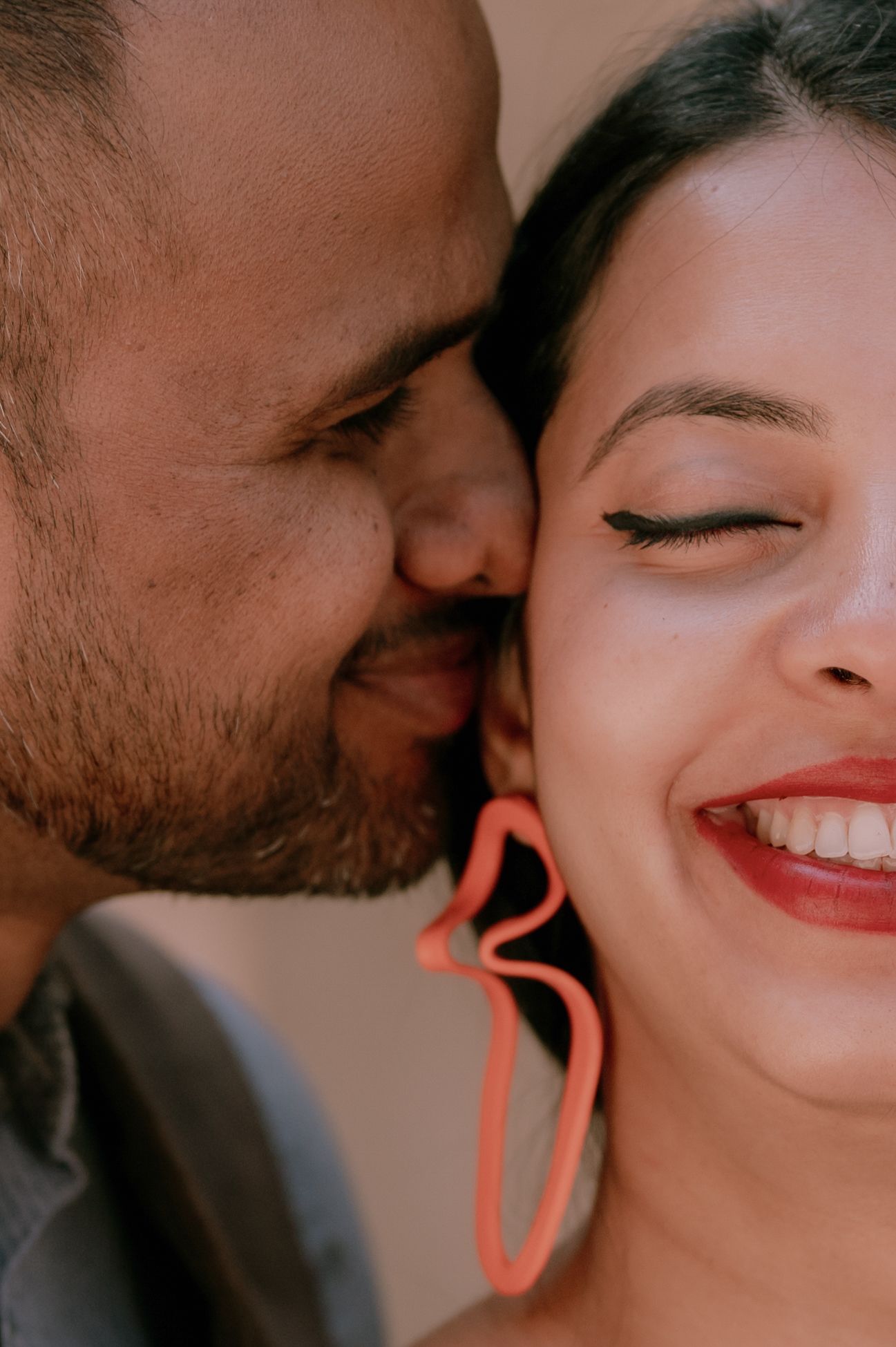 A man with a beard kisses a smiling woman wearing coral heart-shaped earrings on her cheek.