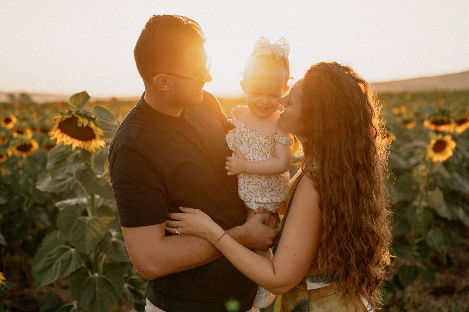 Family with baby girl sharing a tender moment in a sunflower field at golden sunset.