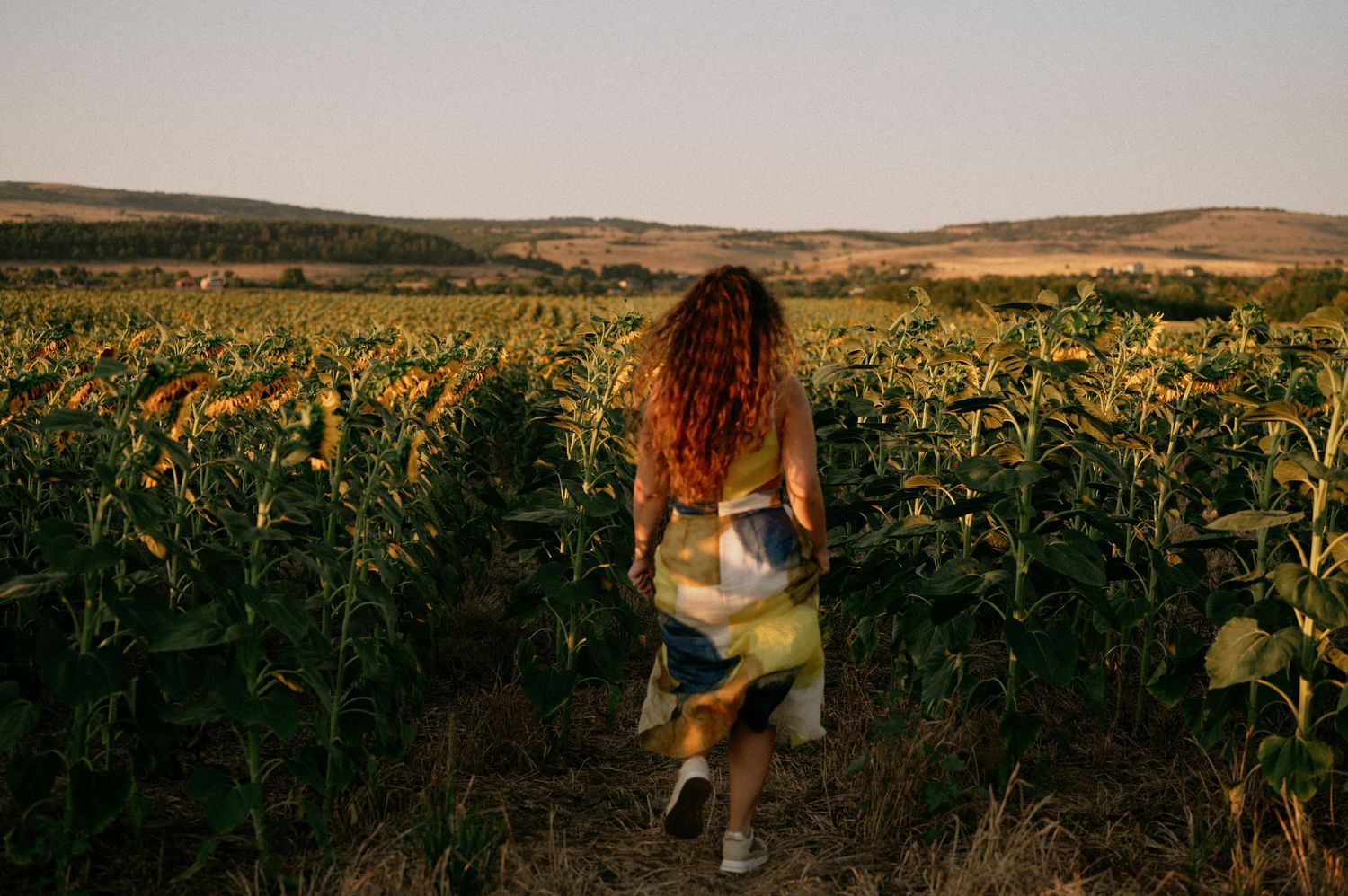 Woman with curly red hair walking through a sunflower field at sunset wearing a flowy dress.