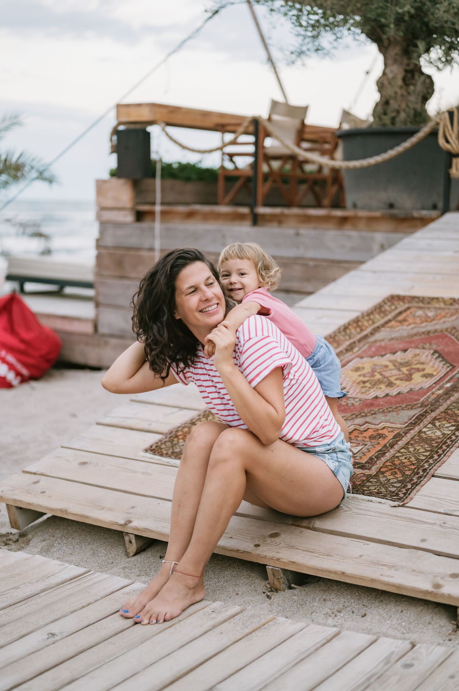 A woman and young child share a joyful hug on wooden beach steps near the ocean.