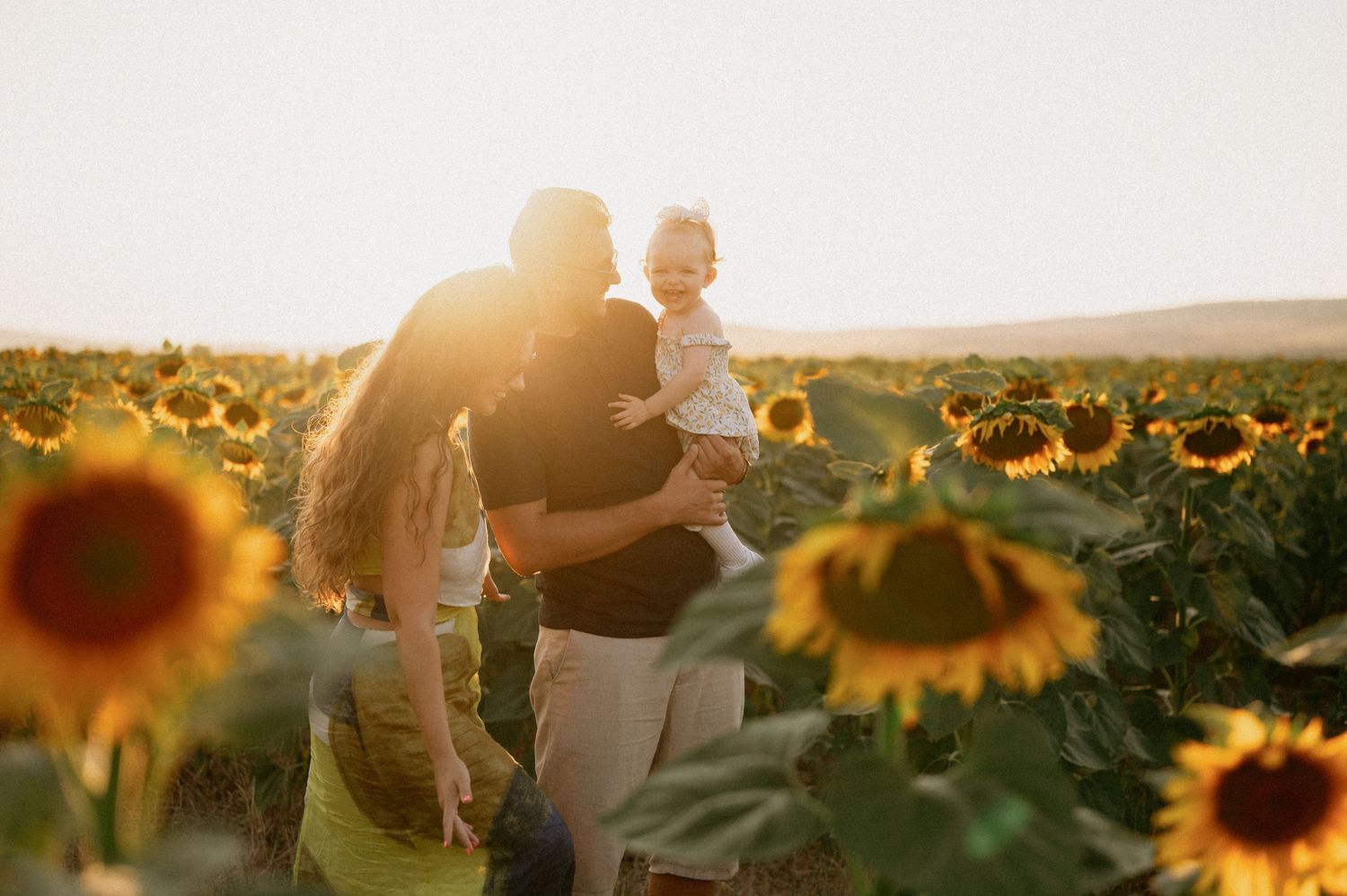 Family with baby enjoying golden hour in a sunflower field, warm sunlight glowing behind them.