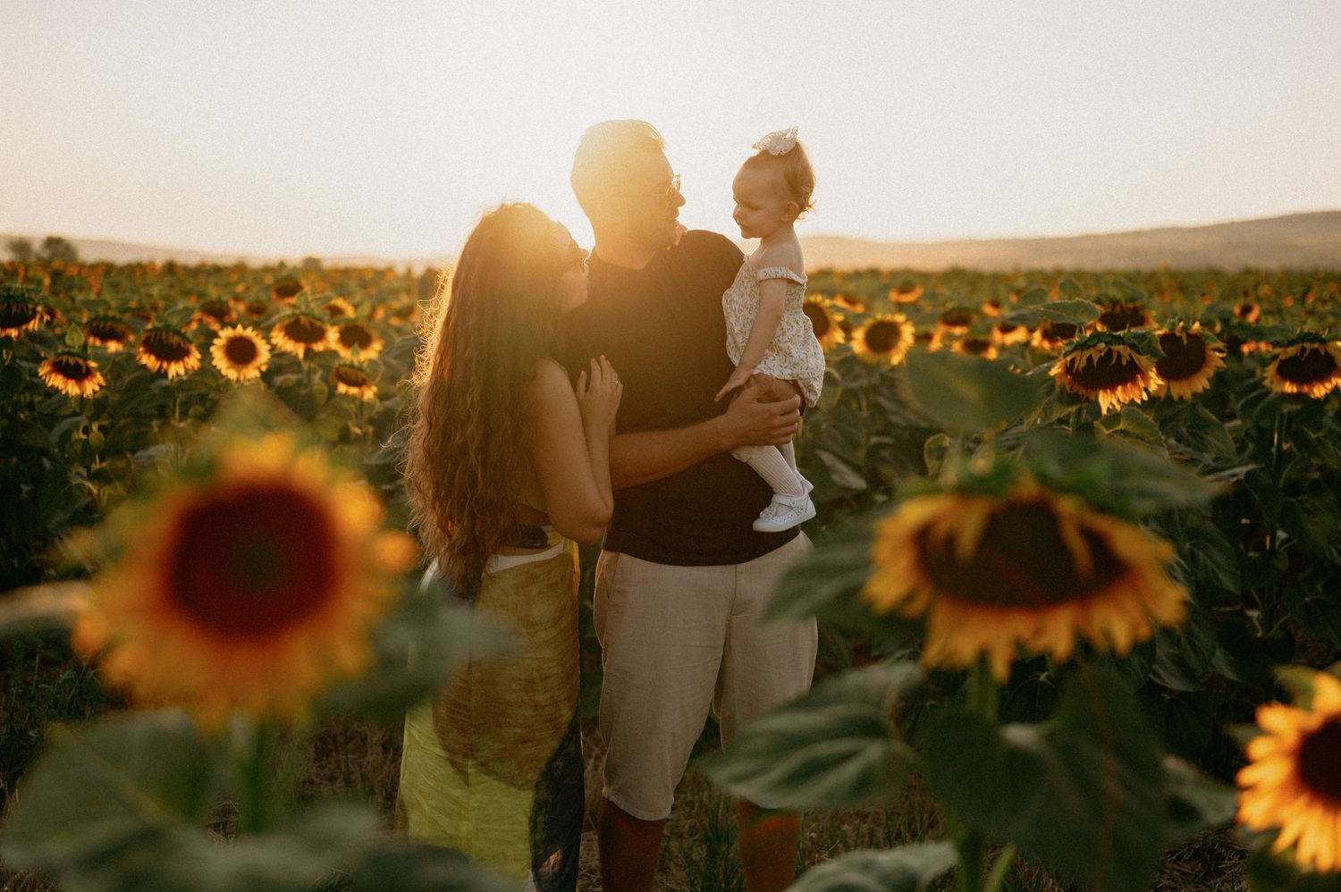 Family with baby posing in a sunflower field at golden hour sunset, warm glowing light illuminating the scene.