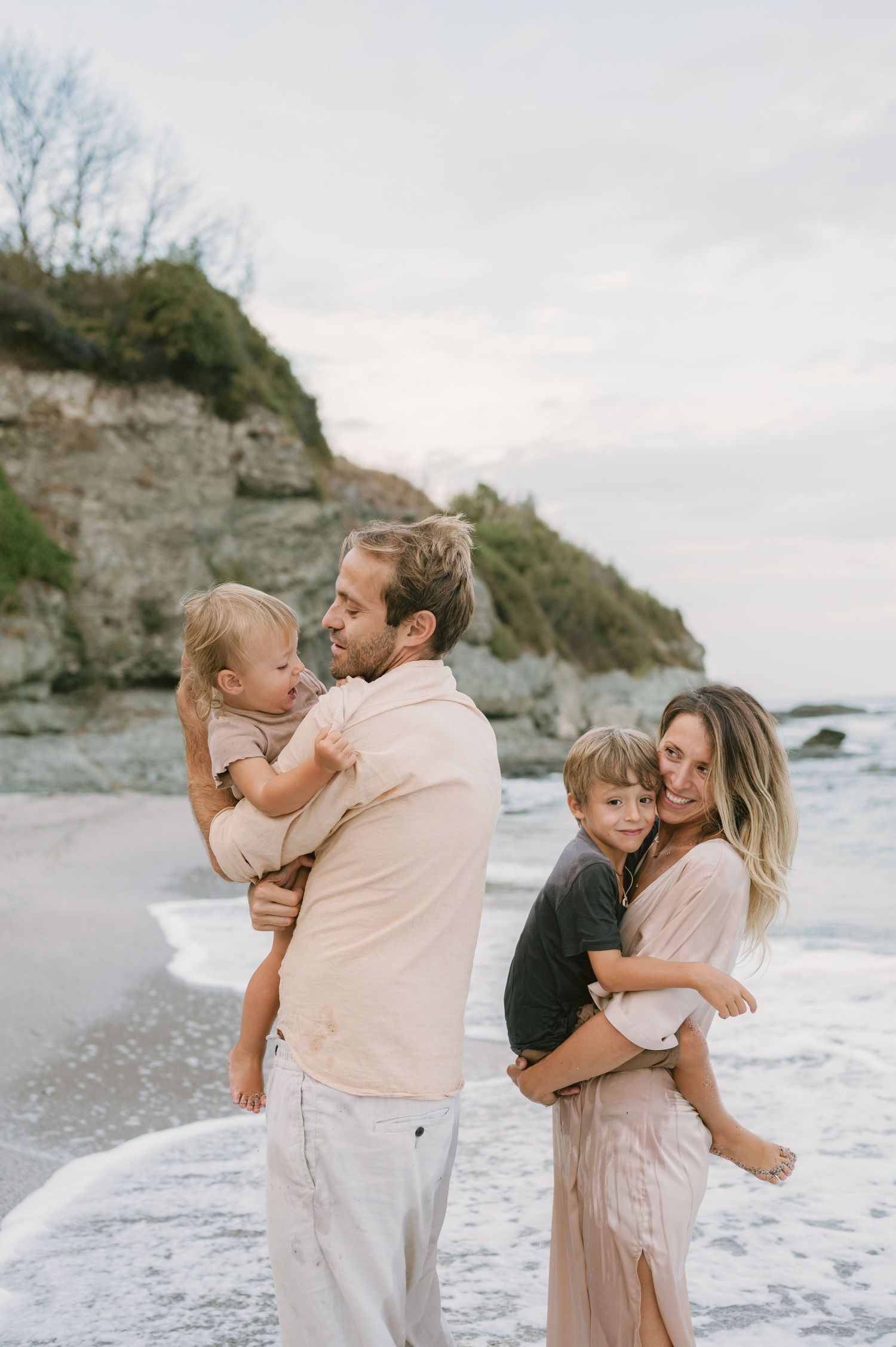 A young family of four poses on a beach, parents holding toddlers with rocky cliffs and ocean waves behind them.