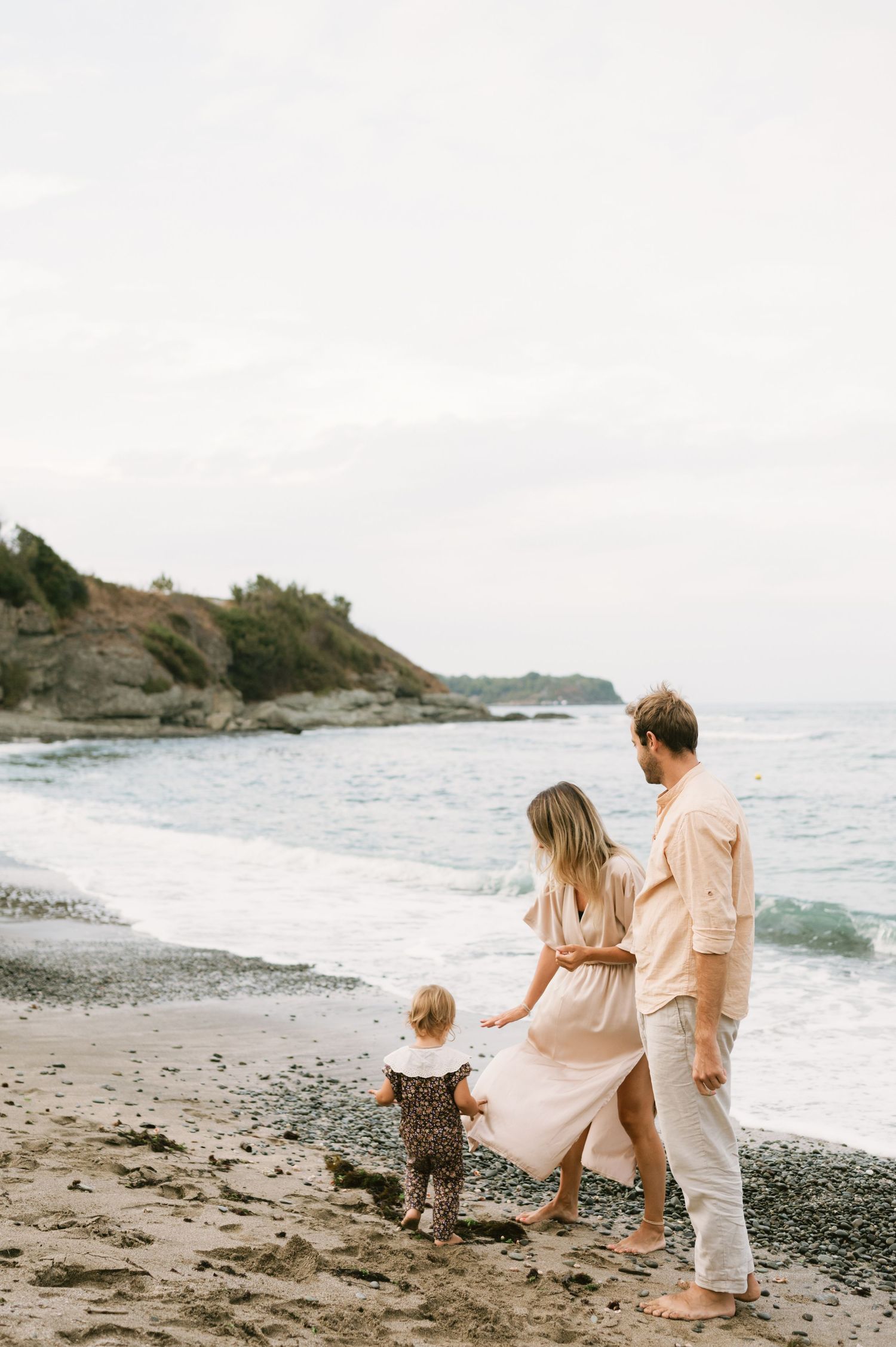 Family of three walking on a rocky beach, parents guiding toddler near ocean waves on an overcast day.