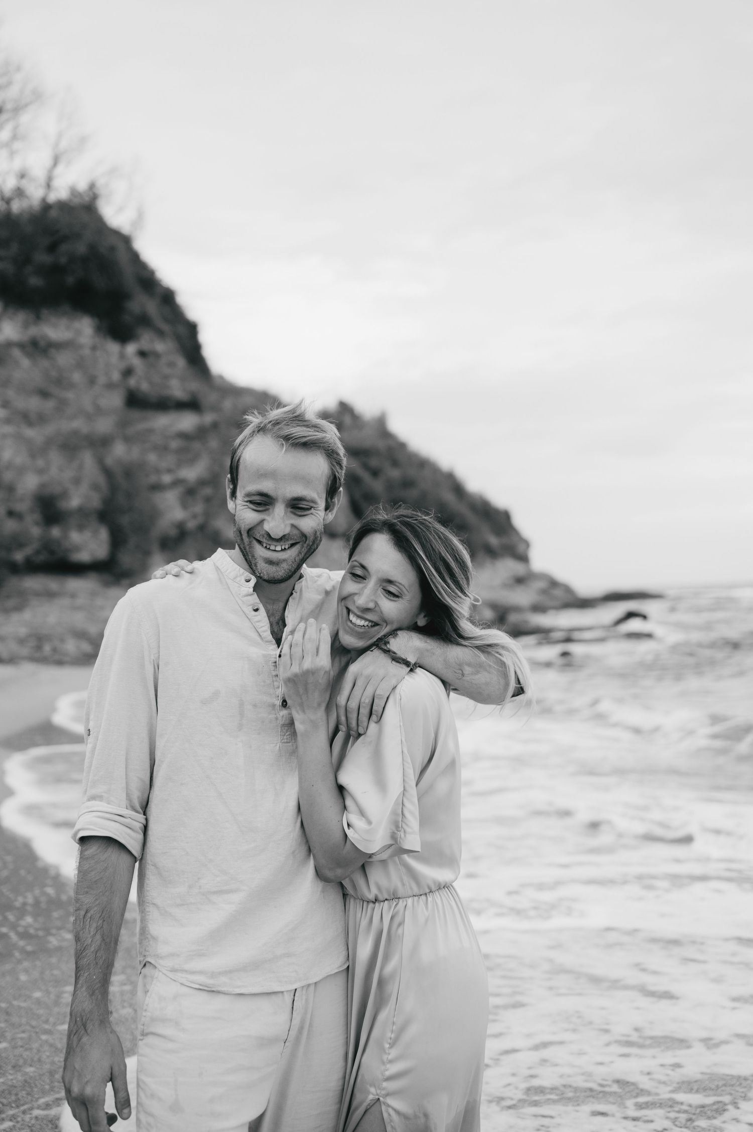 Black and white photo of a happy couple embracing and laughing on a rocky beach shoreline.