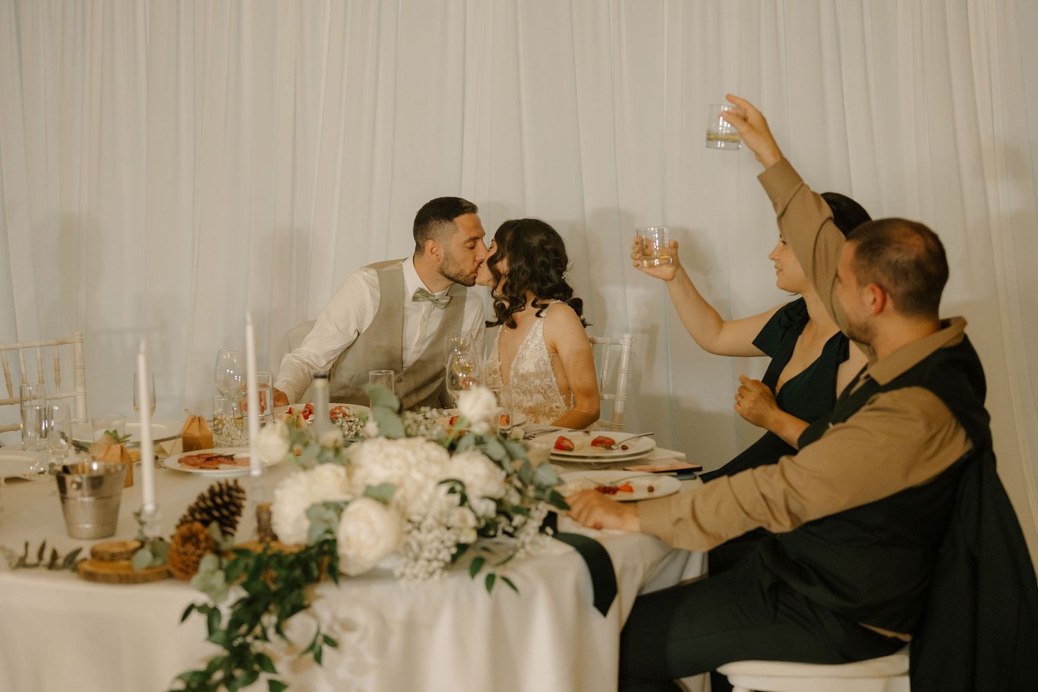 Couple kissing at elegant wedding reception table with white floral centerpiece and candles while guests toast.