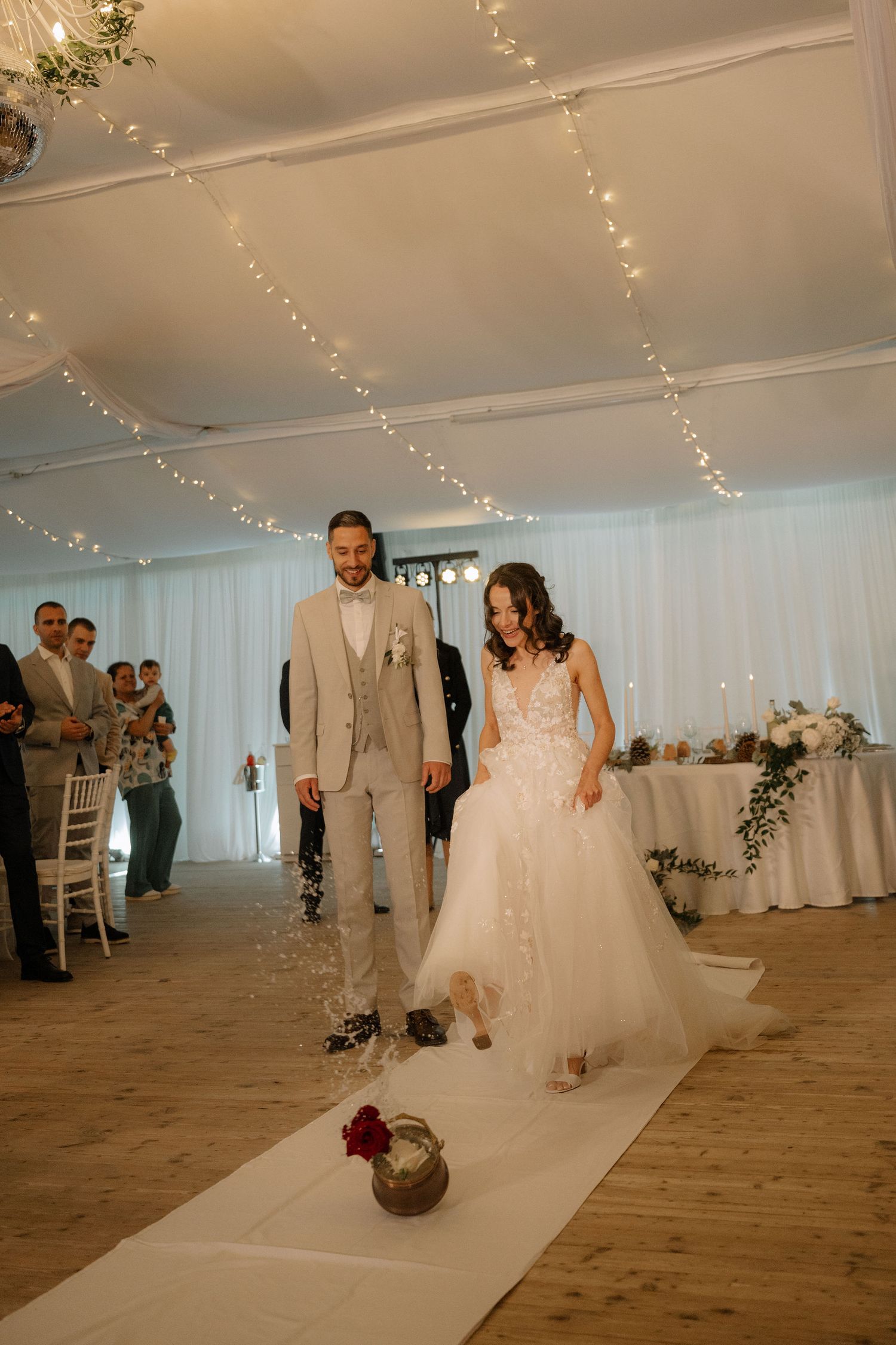 Bride in white tulle gown and groom in beige suit walk down aisle in fairy light decorated wedding venue.