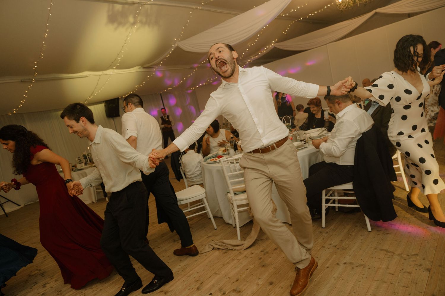 Guests dancing joyfully at a wedding reception in a decorated tent venue with fairy lights.