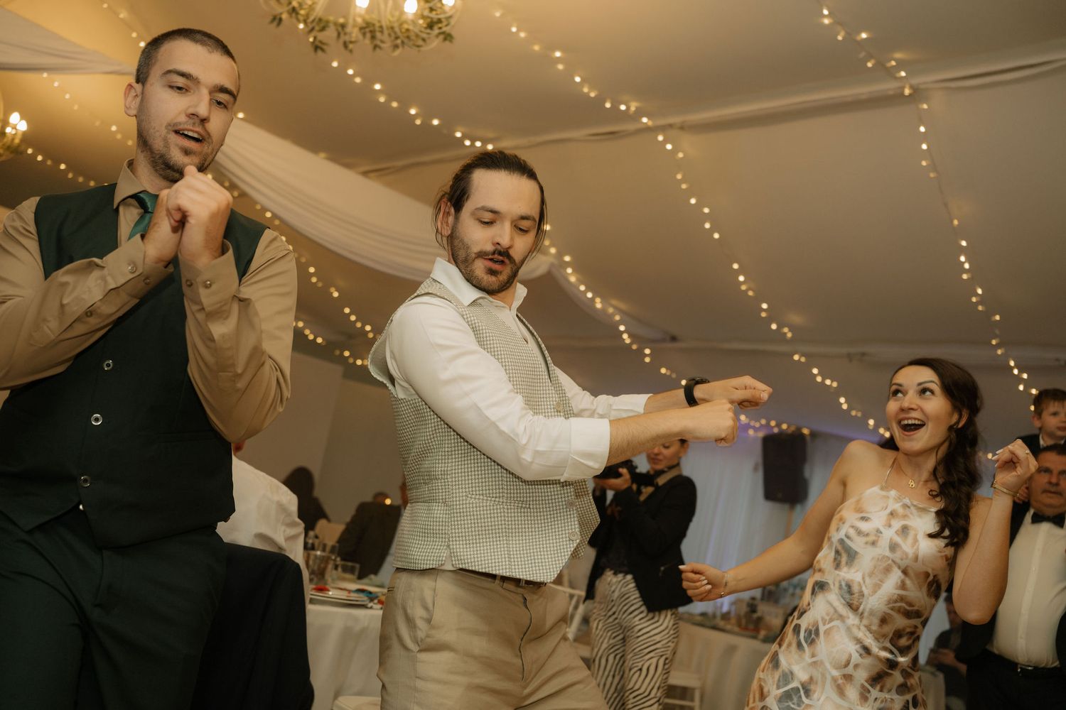 Guests dancing joyfully at a wedding reception under fairy lights in a decorated tent venue.