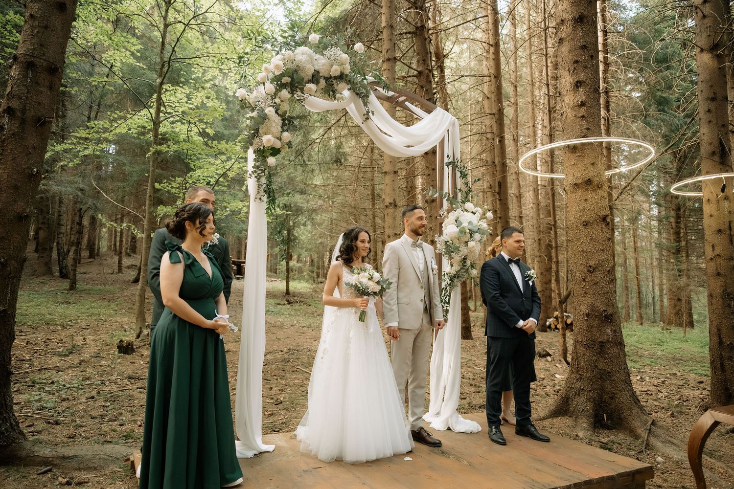 Outdoor forest wedding ceremony with bride and groom under floral arch, flanked by bridesmaid in green and groomsman.