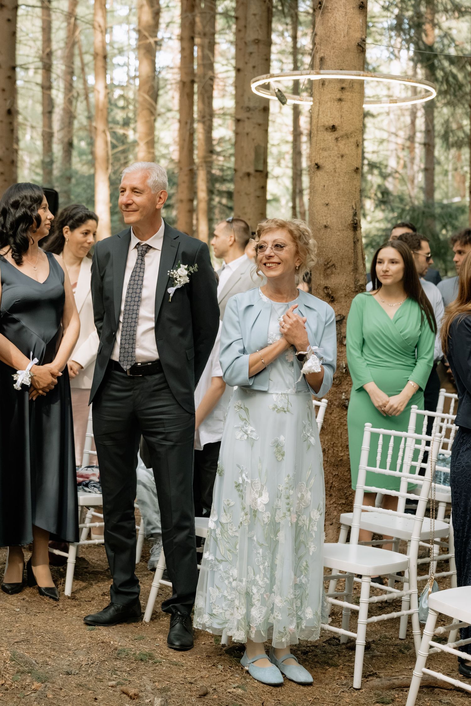 Elegant outdoor wedding ceremony in a forest with guests in formal attire, white chairs, and a hoop decoration overhead.