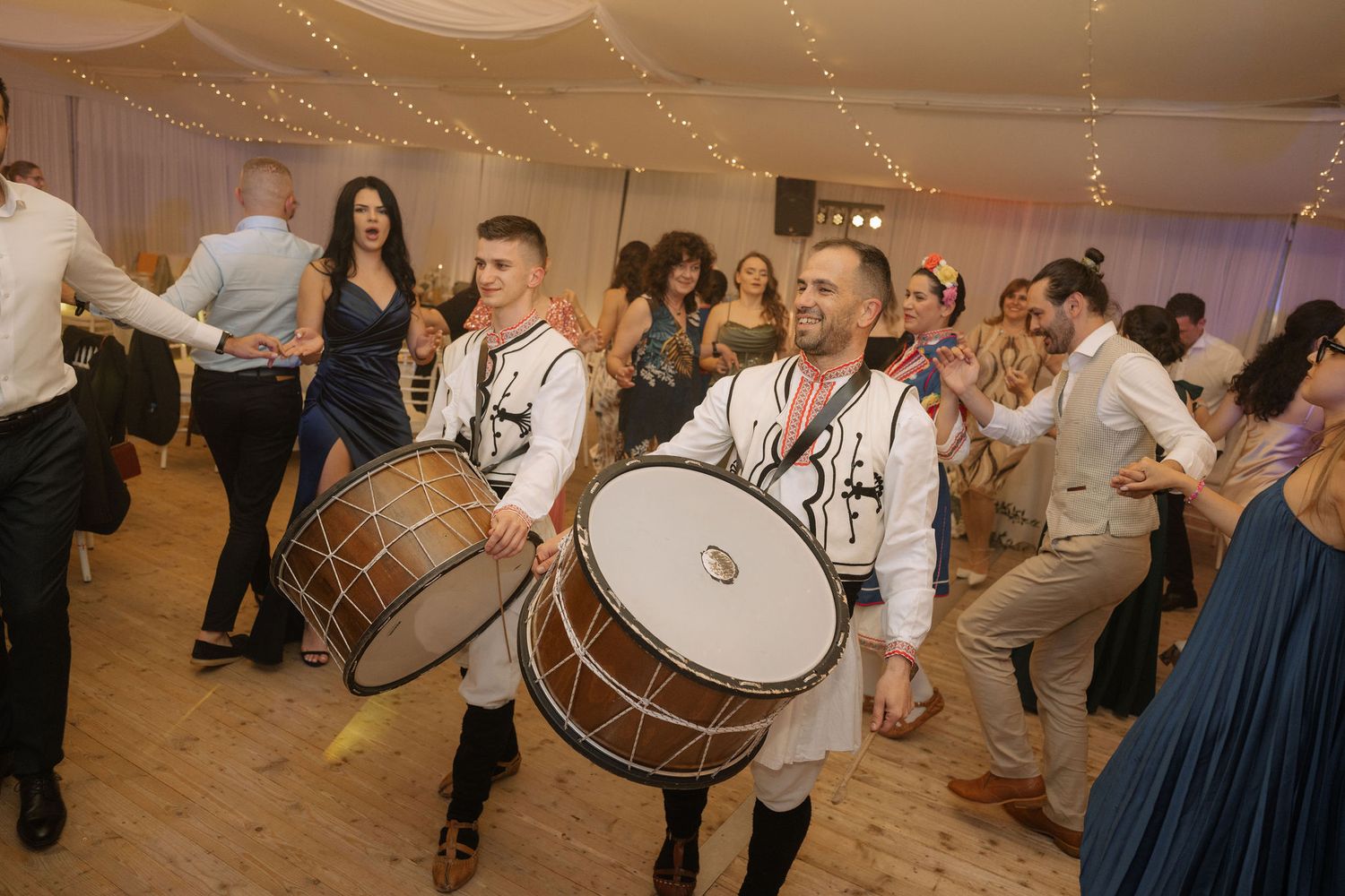 Two drummers in traditional folk costumes perform at a lively wedding reception as guests dance around them.
