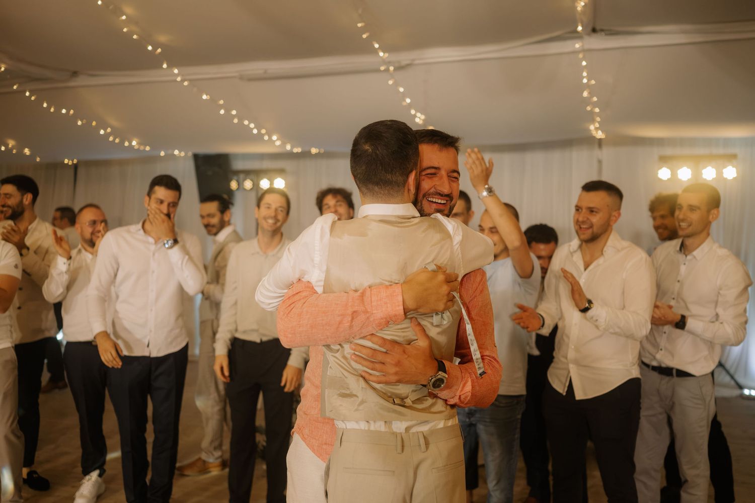 Two grooms share an emotional hug on the dance floor surrounded by smiling guests at a wedding reception.