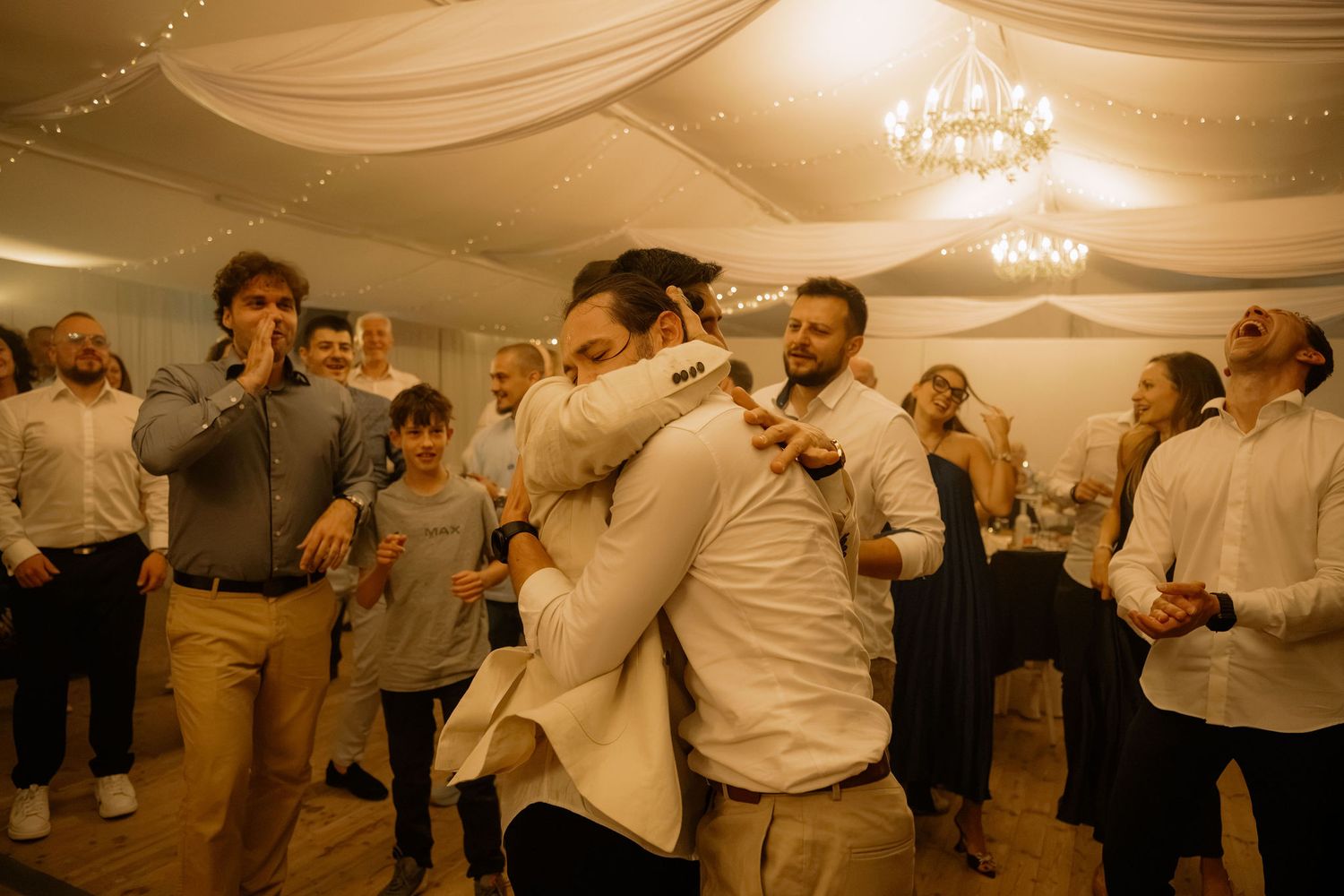 Two men share a warm hug on a wedding dance floor surrounded by celebrating guests in a decorated venue.