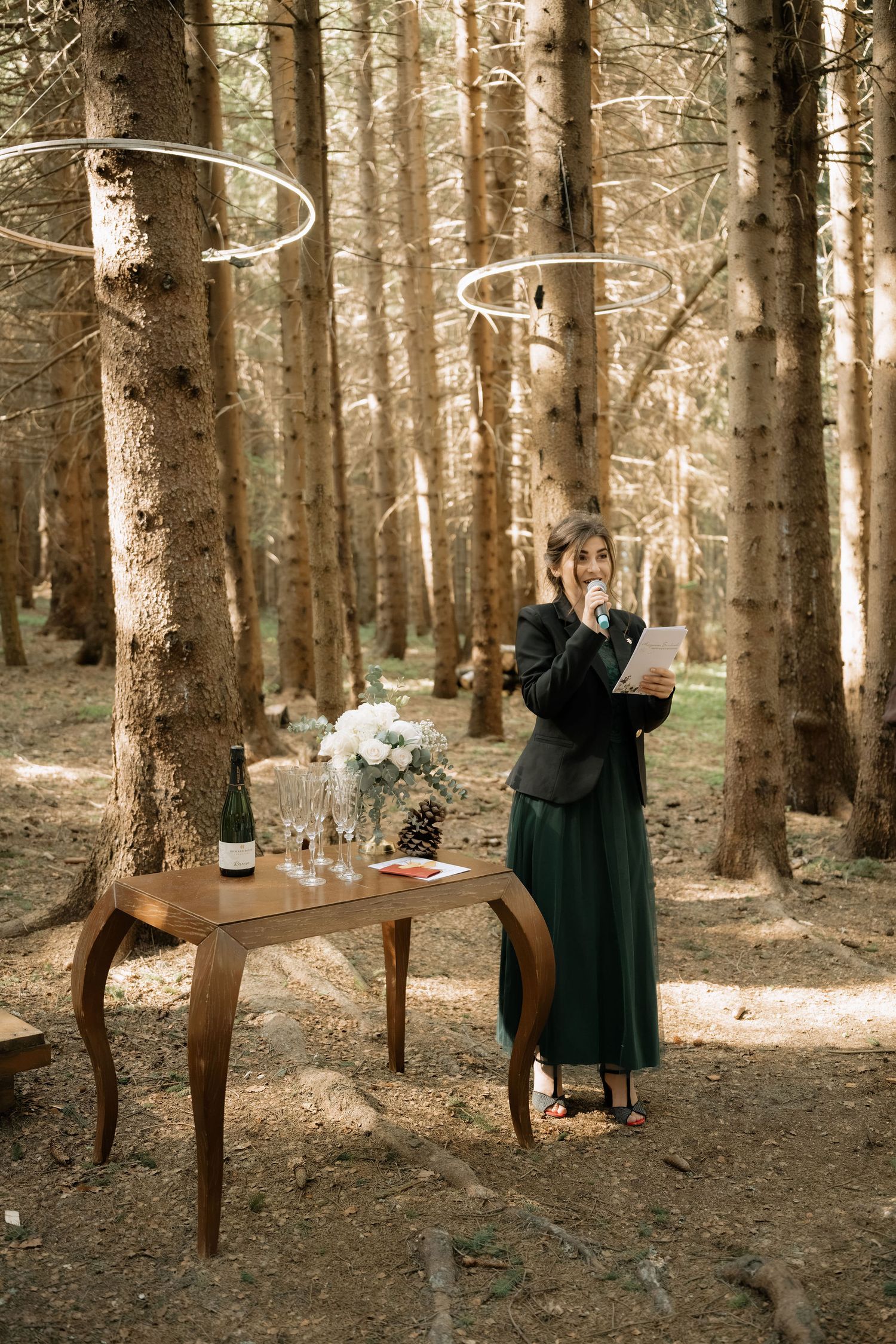 A woman in a green skirt speaks at an outdoor forest wedding ceremony beside a wooden table with flowers and a wine bottle.