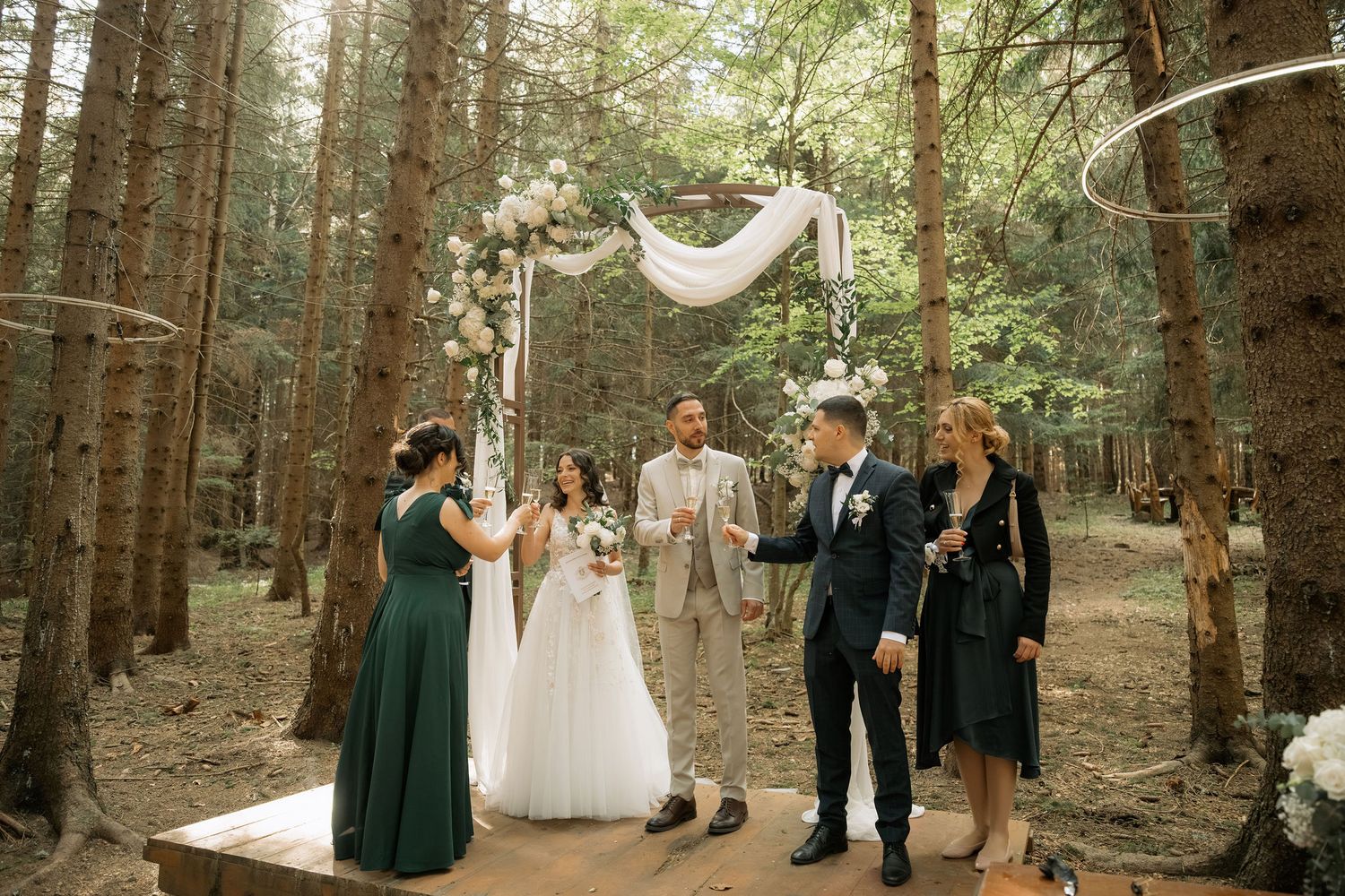 Wedding ceremony in a forest with bride, groom, officiant, and guests standing under a floral arch adorned with white draping.