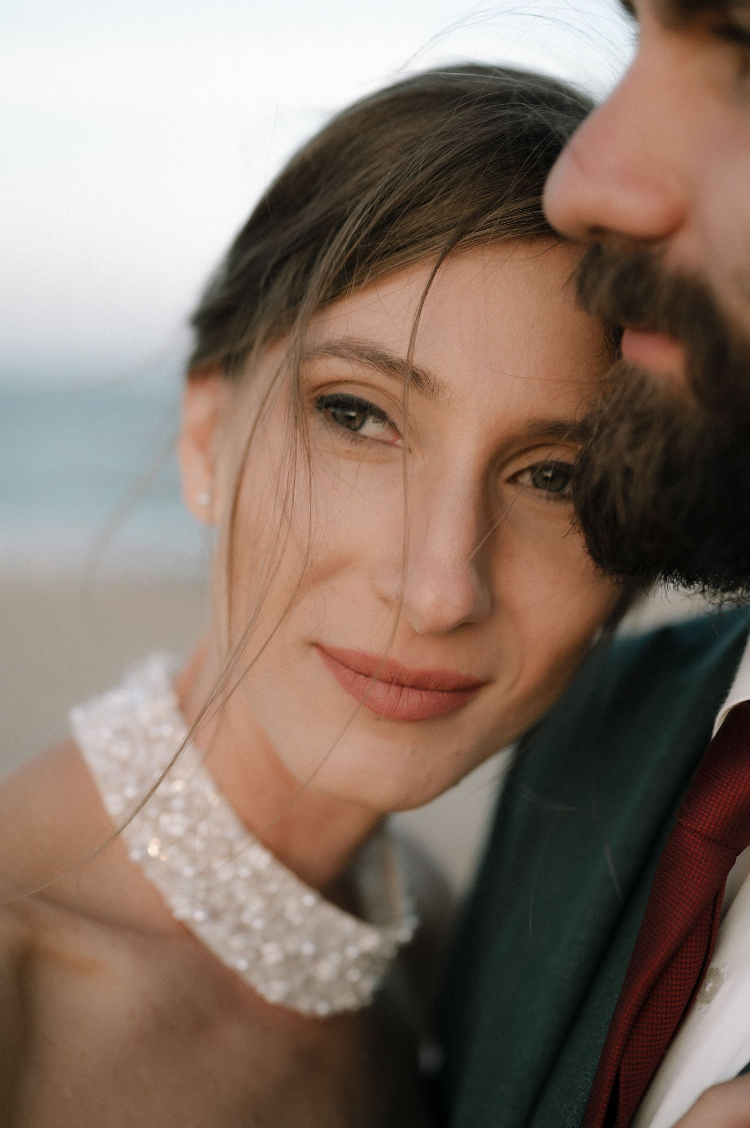 Close-up portrait of a bride leaning against a bearded groom near the ocean, wearing a beaded choker necklace.