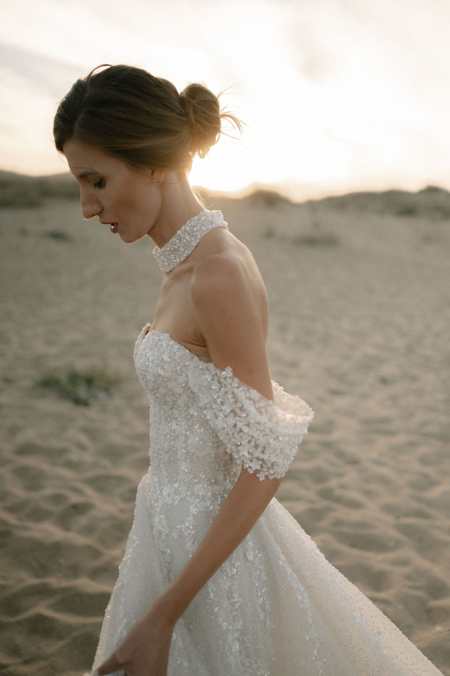 Bride in off-shoulder lace halter wedding gown poses on sandy desert dunes at golden sunset.