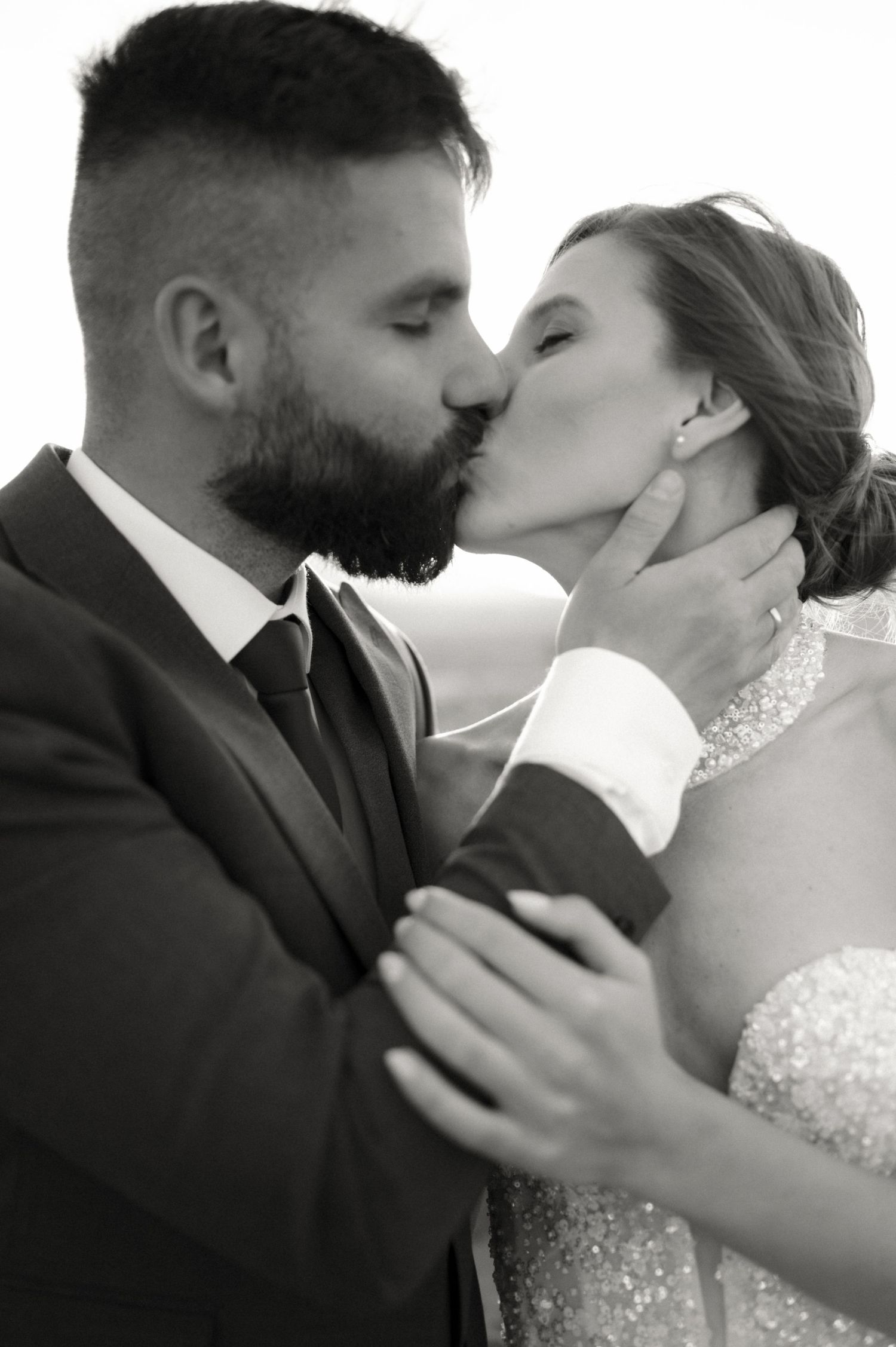 Black and white wedding photo of bride and groom sharing a romantic kiss, groom in suit, bride in beaded gown.