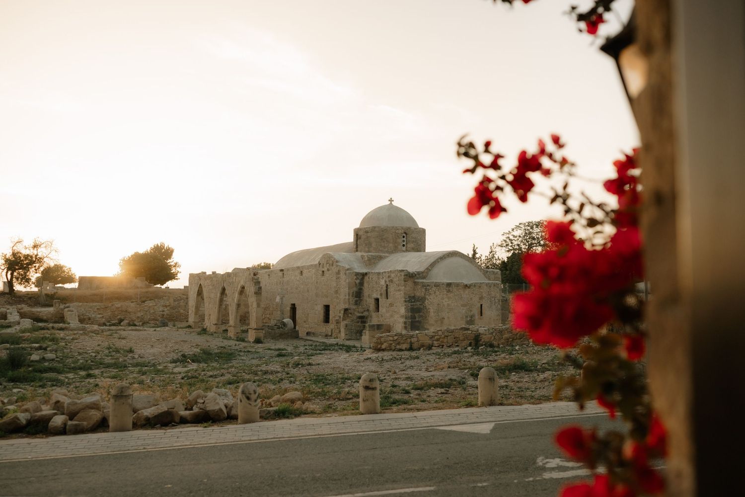 Ancient stone church with a dome glows at sunset, framed by vibrant red bougainvillea flowers in Cyprus.