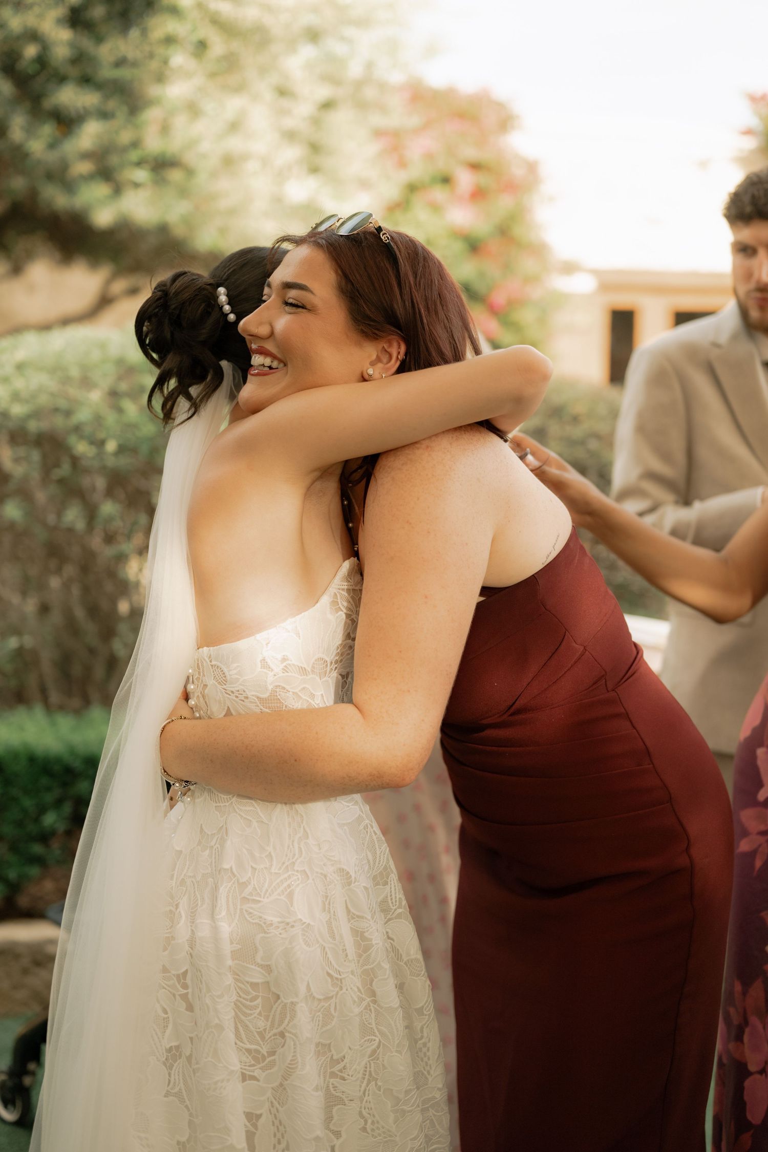 Bride in strapless lace gown and veil sharing joyful hug with bridesmaid in burgundy dress at outdoor wedding.