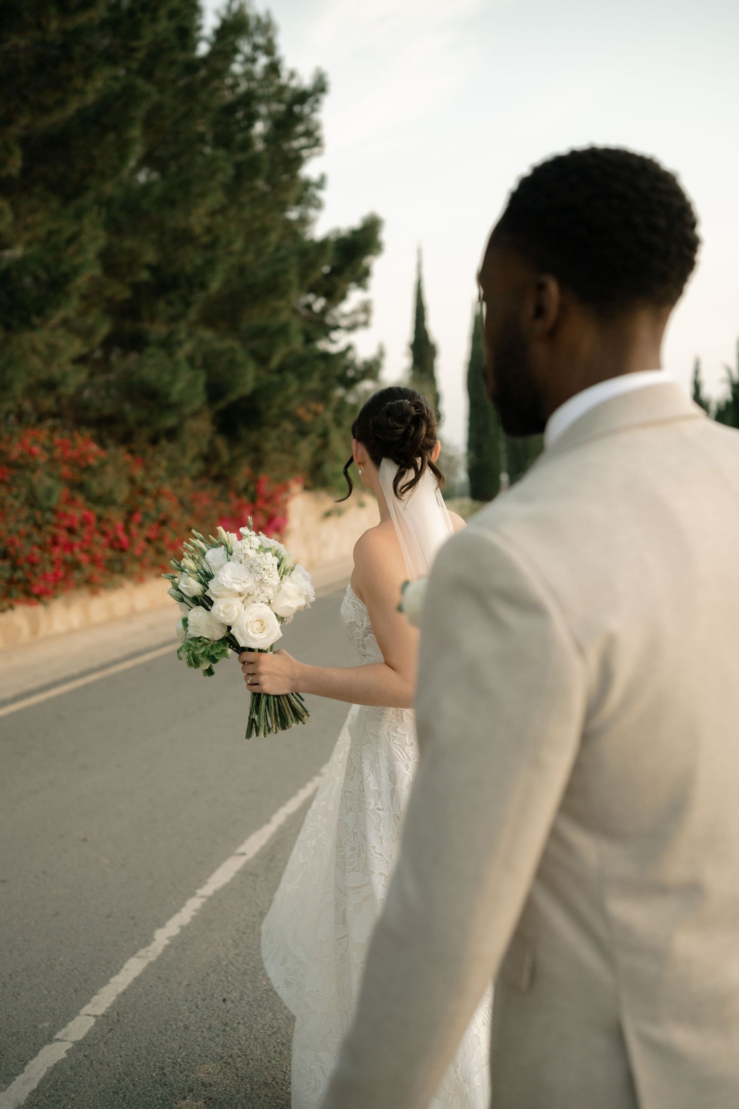Interracial couple walking on road, bride holding white bouquet in wedding dress, groom in beige suit, cypress trees.