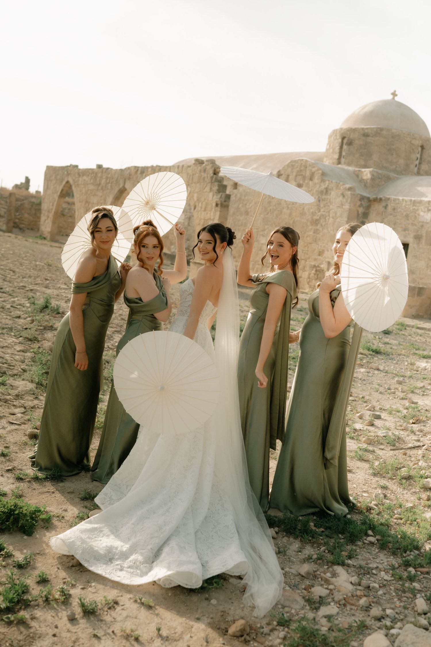 Bride in white gown with bridesmaids in sage green dresses holding white parasols at ancient stone ruins.