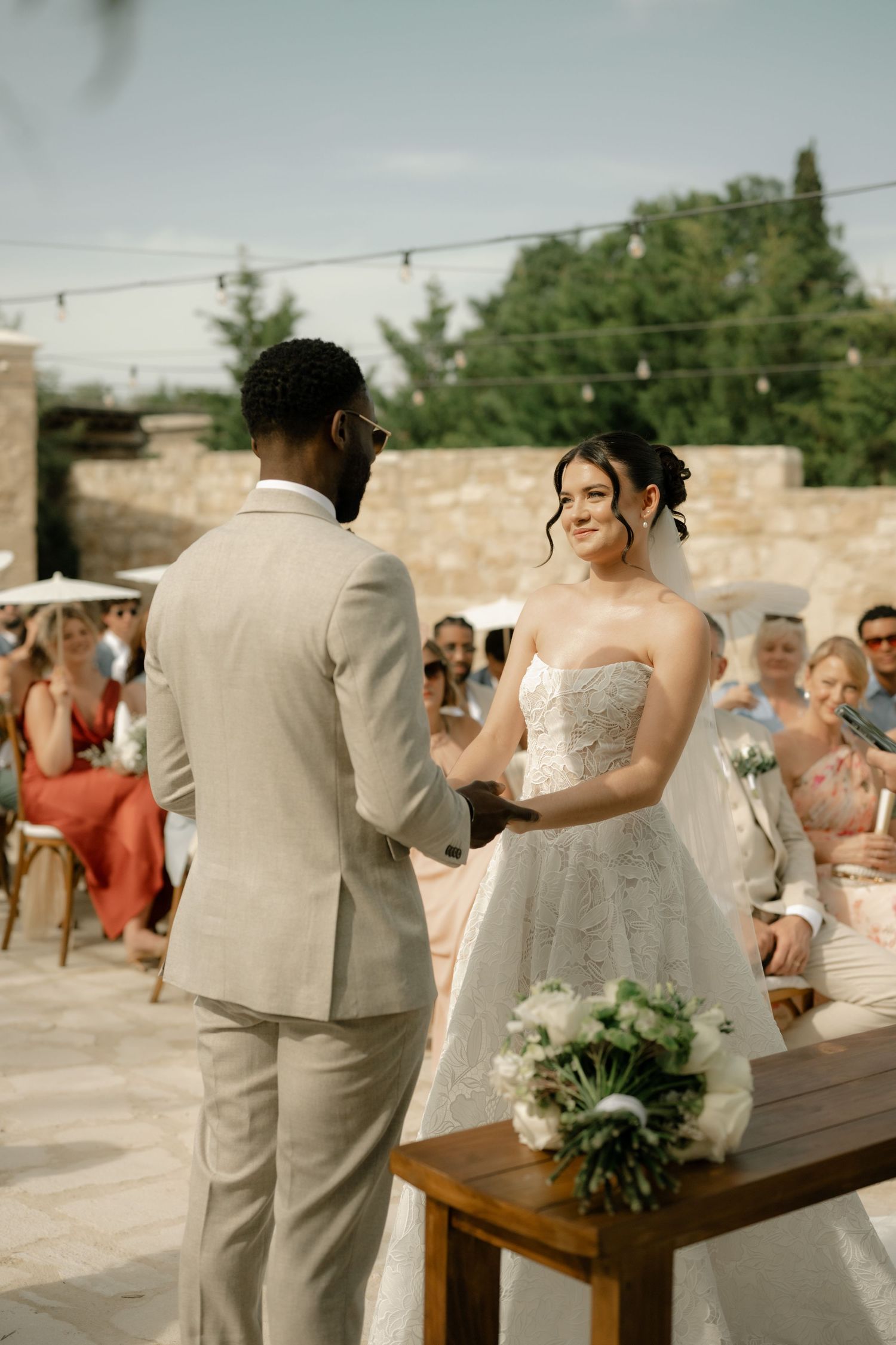 Bride and groom exchanging vows at an outdoor wedding ceremony with seated guests in the background.