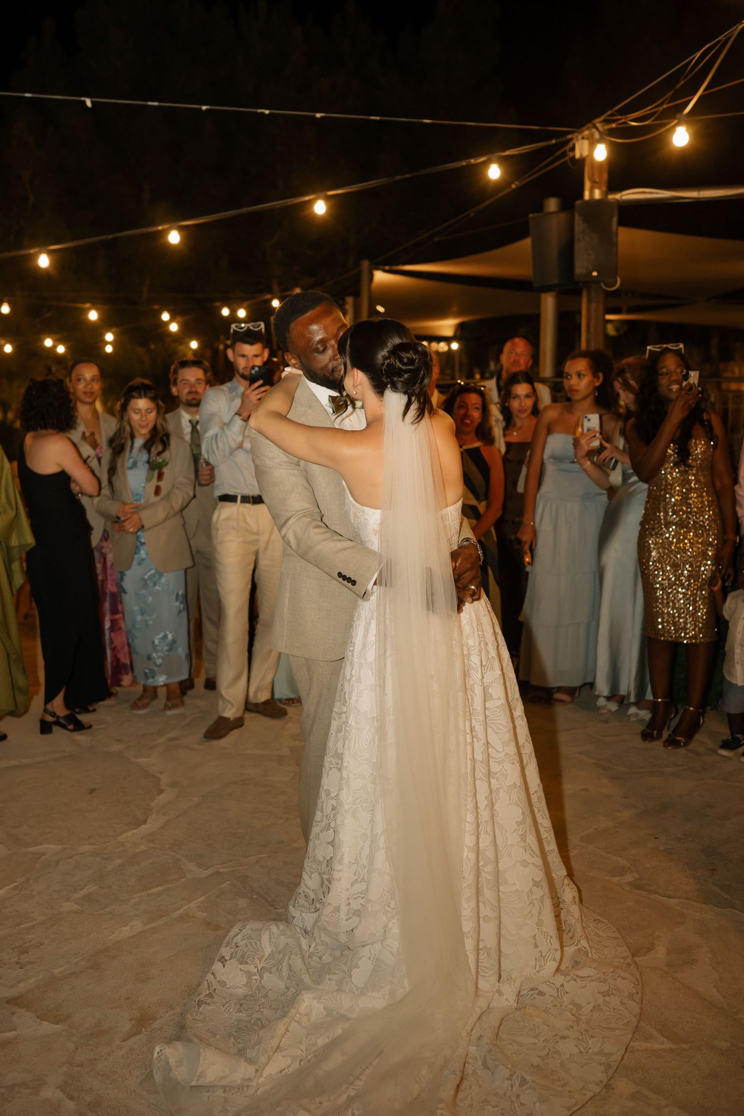 Bride and groom share first dance under string lights at outdoor wedding reception with guests watching.