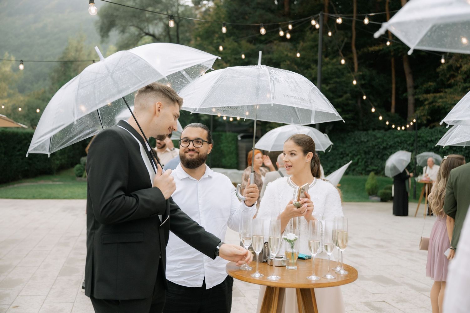 A group of wedding guests gather under clear umbrellas during a rainy outdoor celebration.