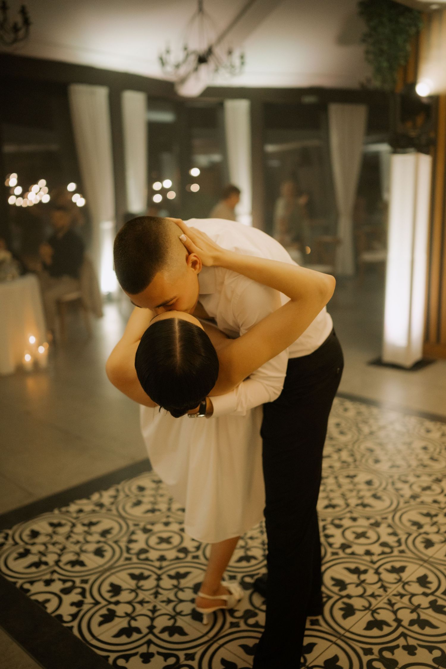 A romantic couple dances together on a black and white patterned floor in a dimly lit ballroom setting.