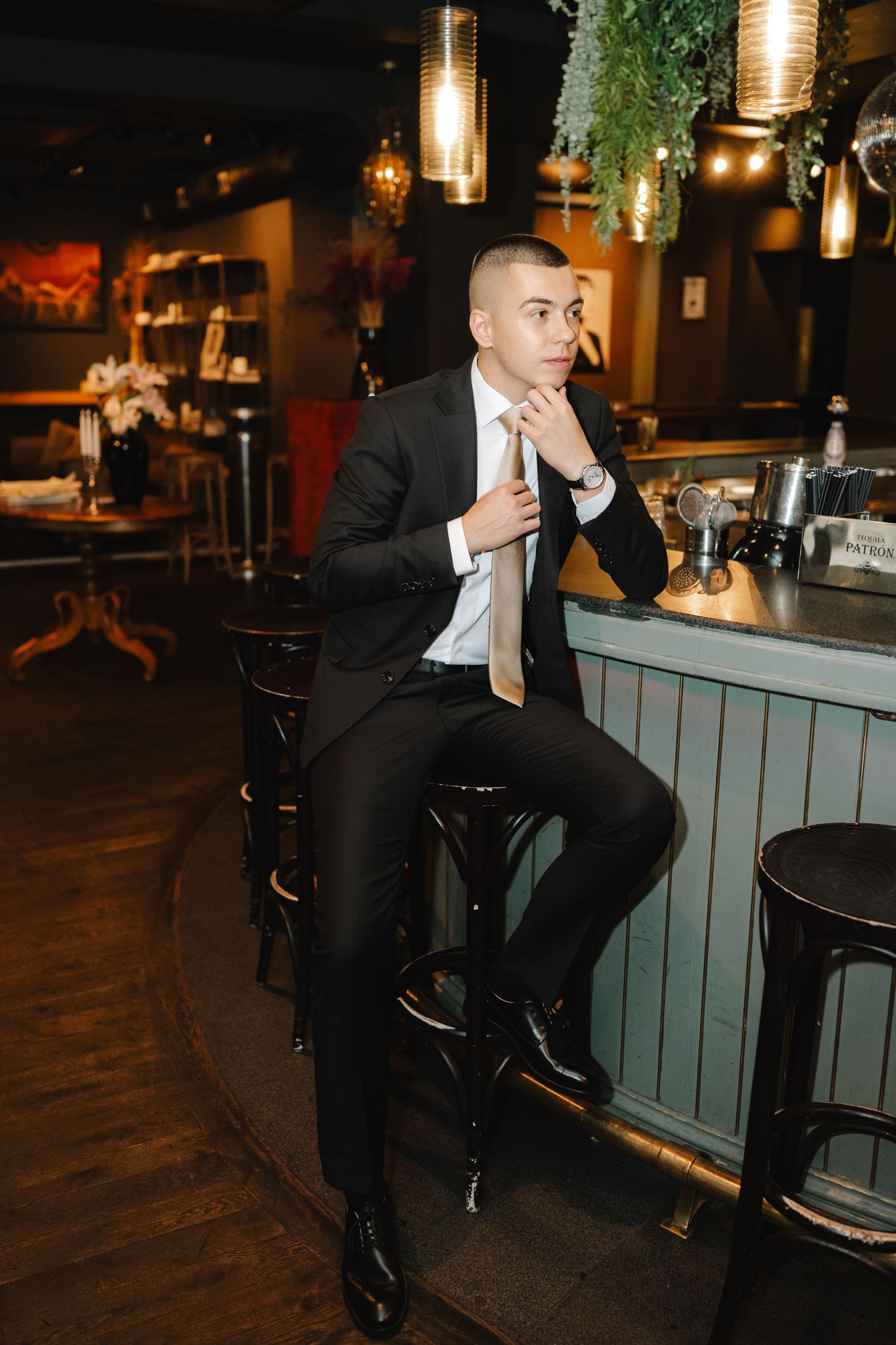 A well-dressed person in a black suit sits thoughtfully at an upscale bar with ambient lighting and hanging pendant lamps.