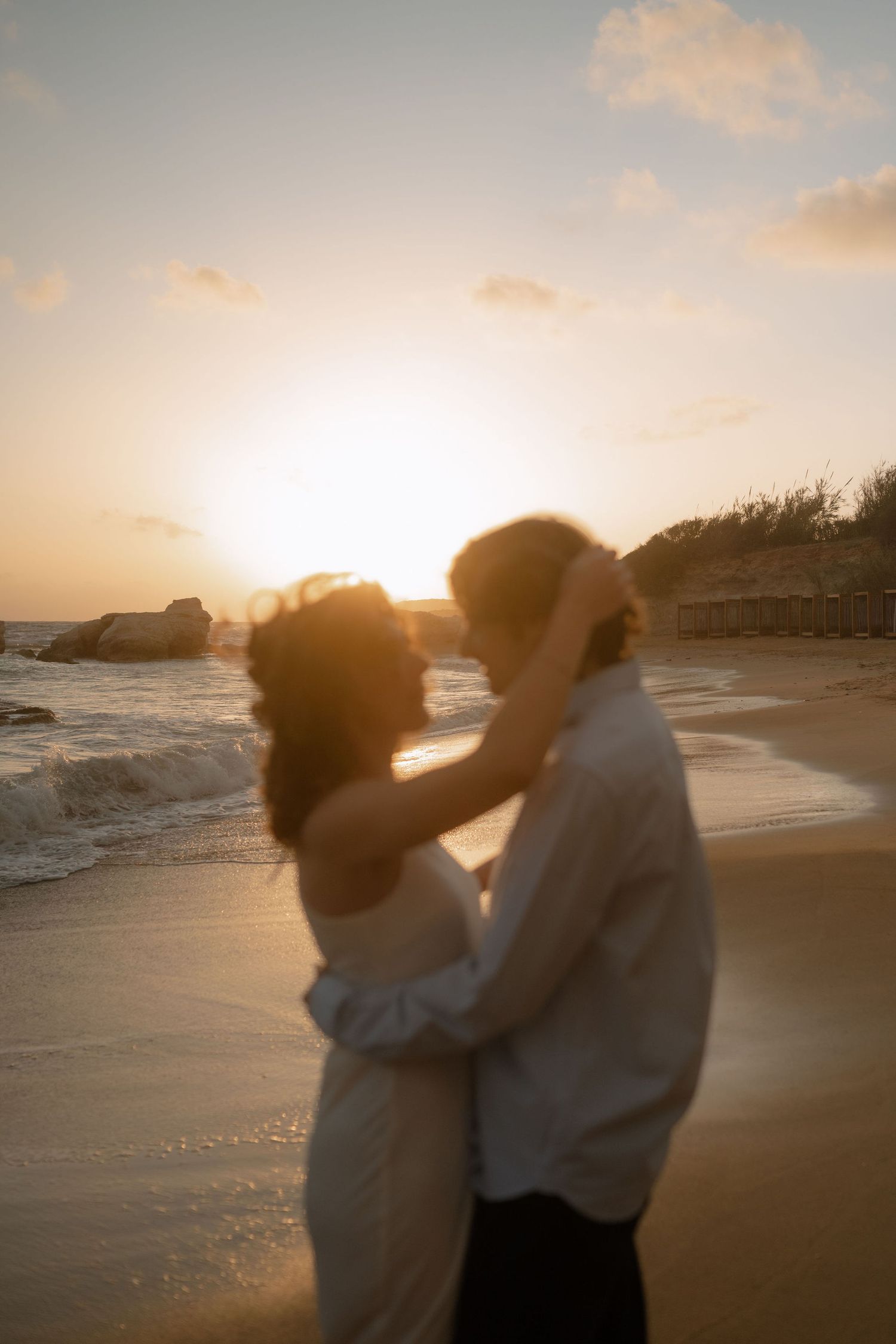 Couple embracing on a sandy beach at golden sunset, waves crashing behind them in a romantic silhouette.
