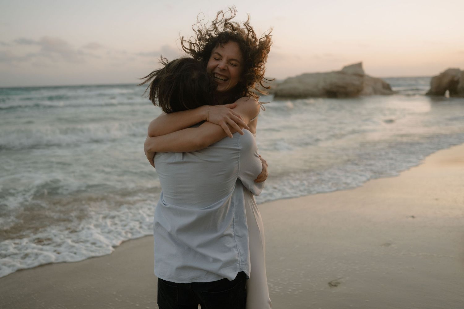 Couple embracing on a sandy beach at sunset with ocean waves and rocky cliffs in the background.