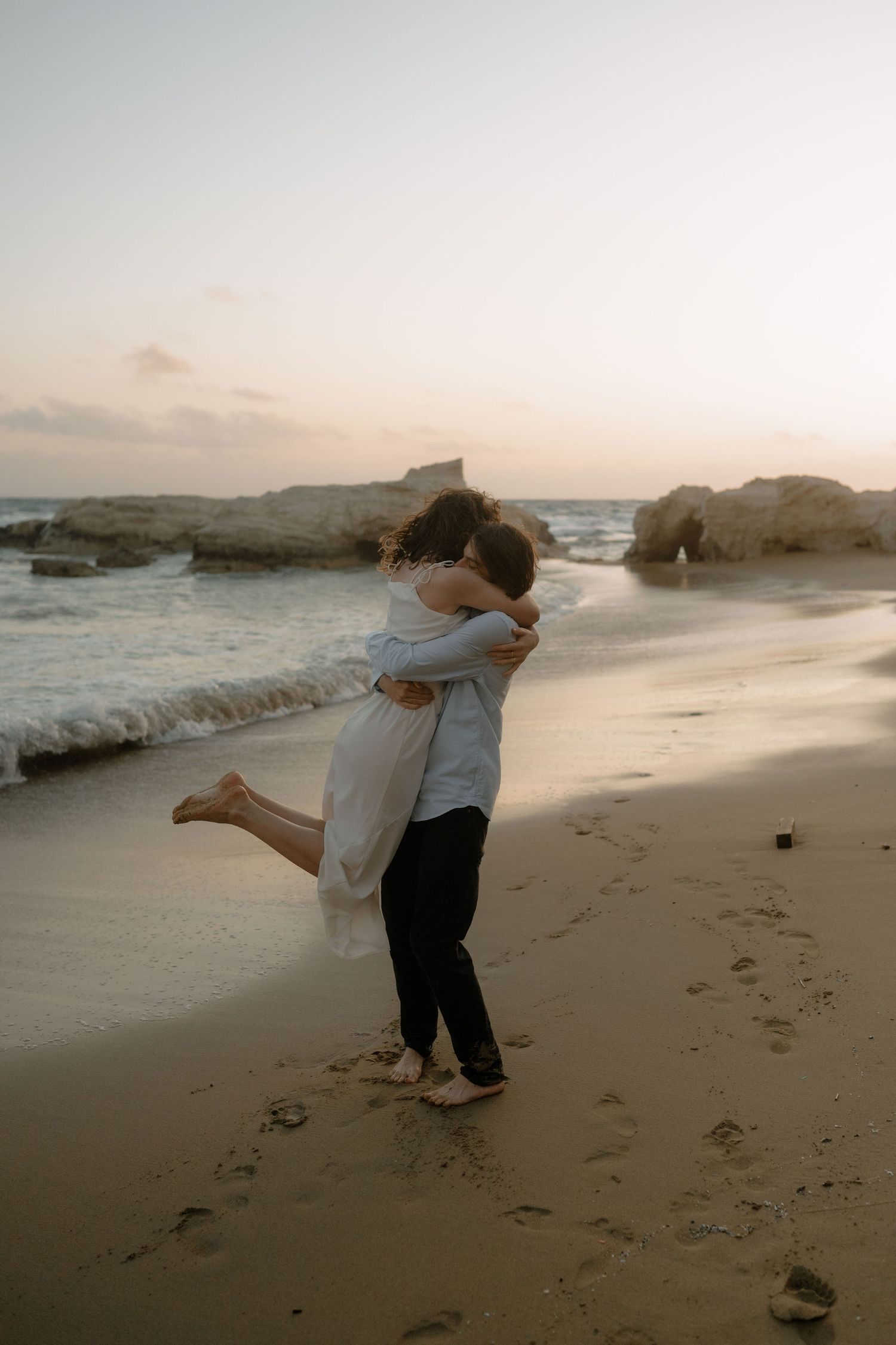 Couple embracing on a sandy beach at sunset with ocean waves and rocky cliffs in the background.